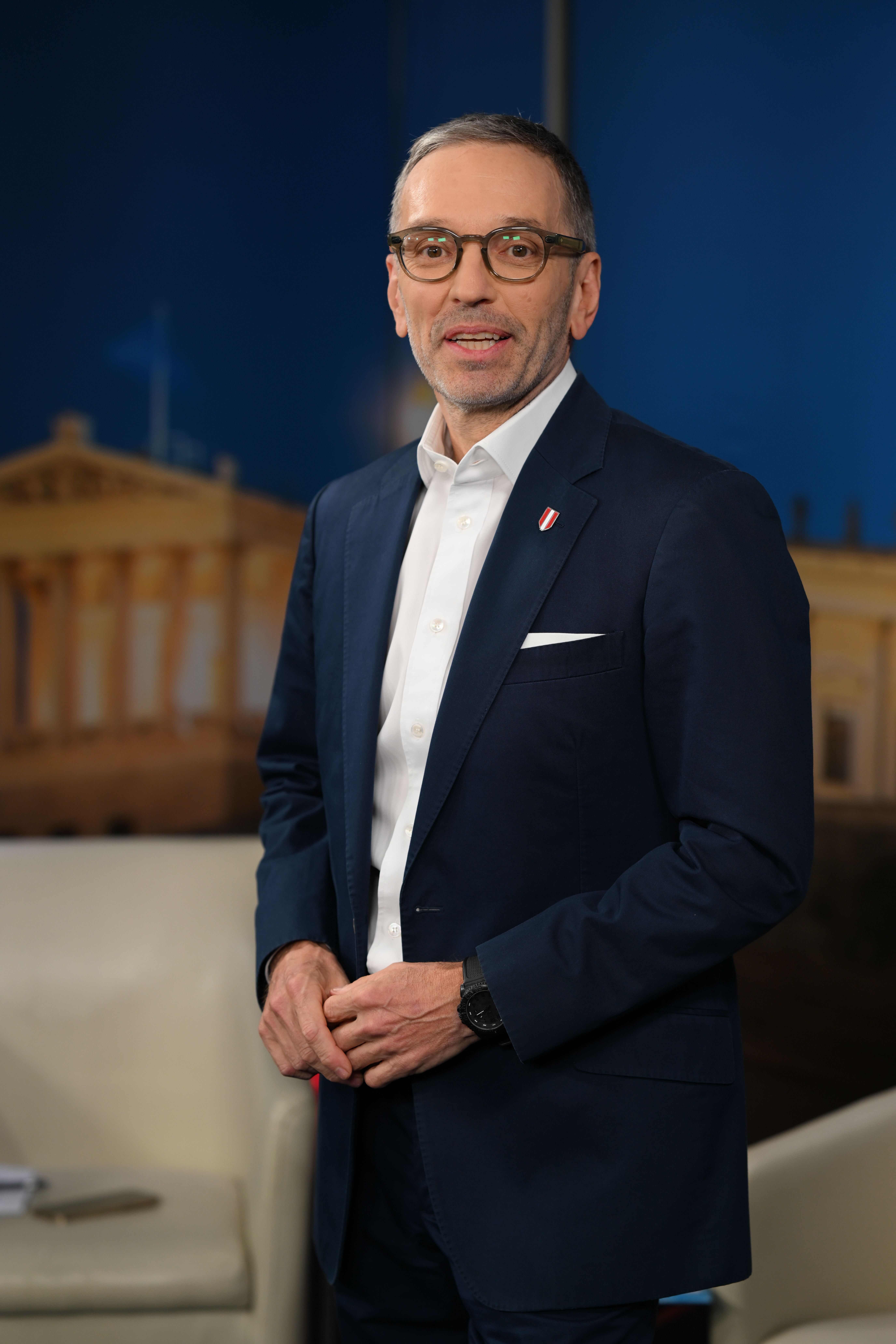 Ein Mann mit Brille im blauen Anzug steht vor einem unscharfen Hintergrund.