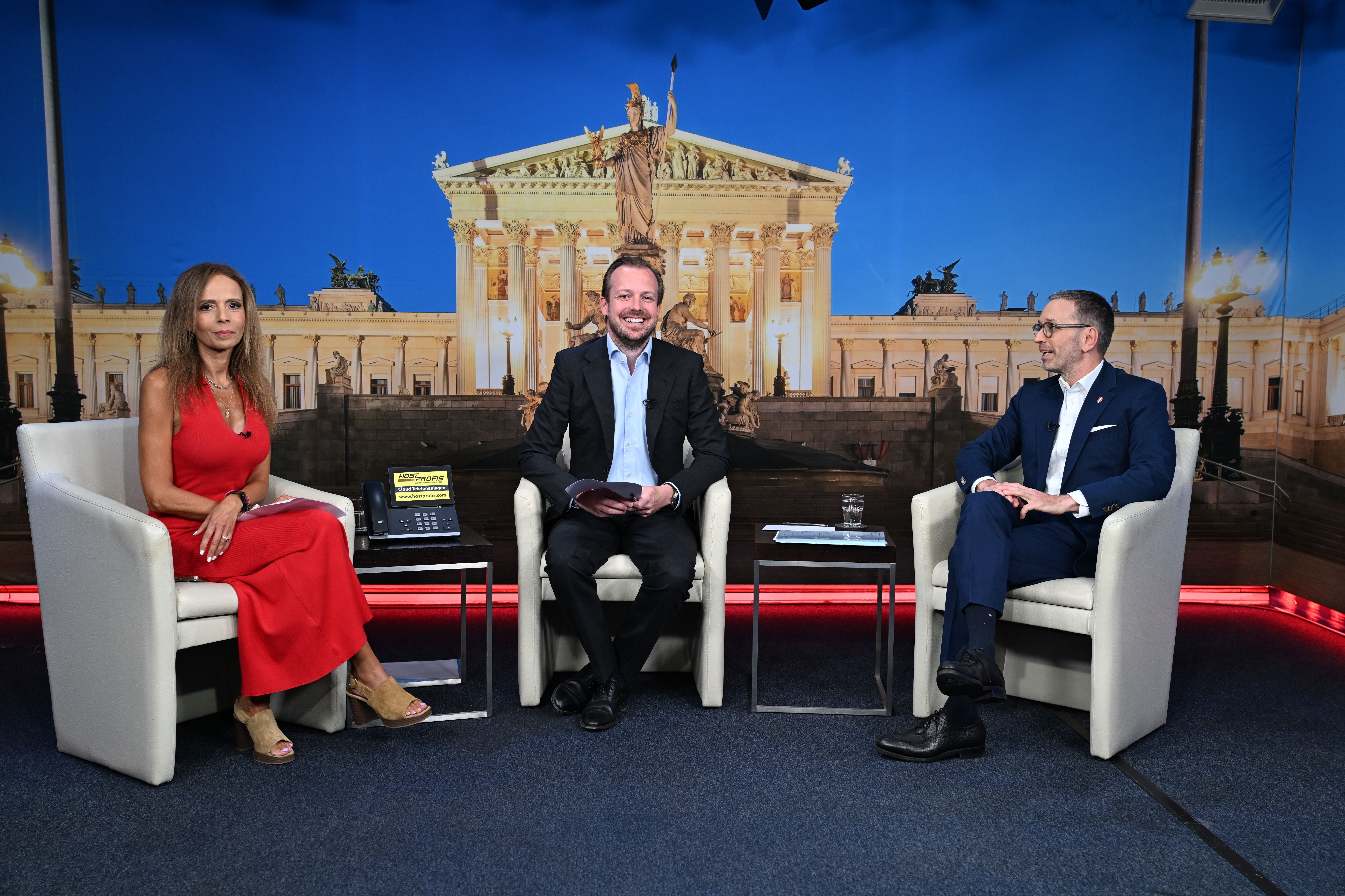 Drei Personen sitzen in einem Fernsehstudio vor einem Bild des österreichischen Parlaments.