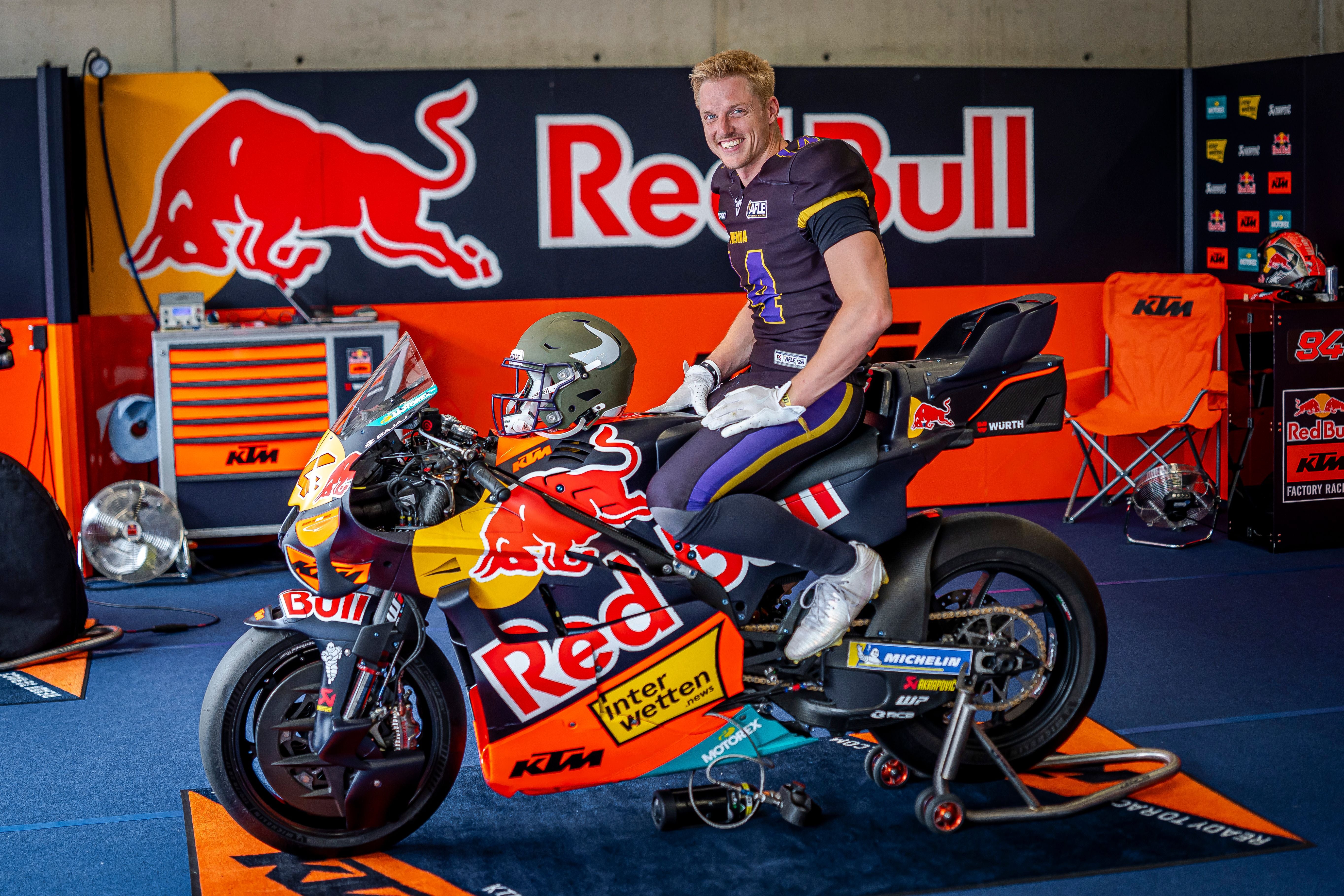 Ein Mann in Footballkleidung sitzt auf einem KTM-Red Bull Rennmotorrad in einer Box.