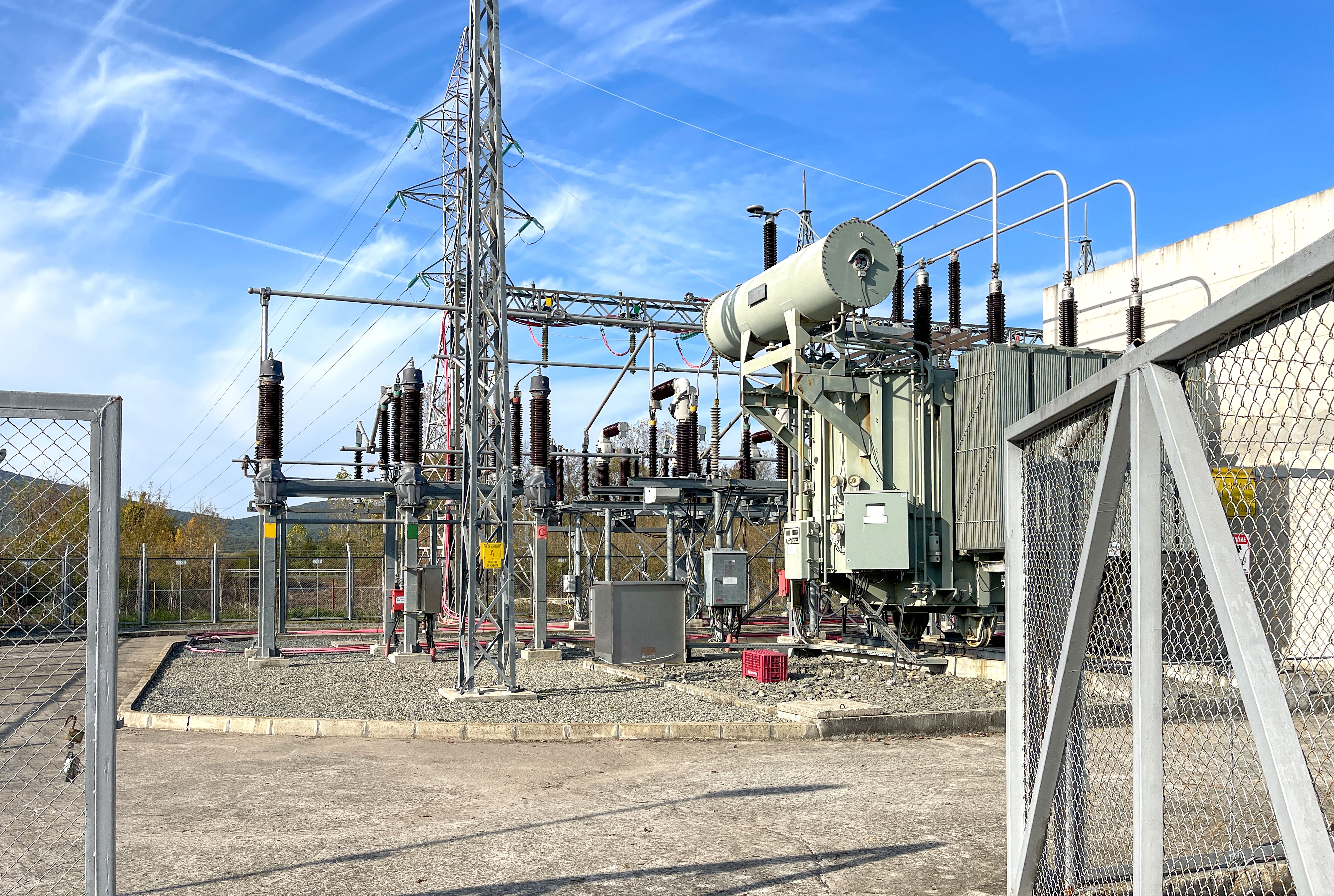 Elektrisches Umspannwerk mit Transformator, Hochspannungsisolatoren und Übertragungsmast bei Tageslicht.