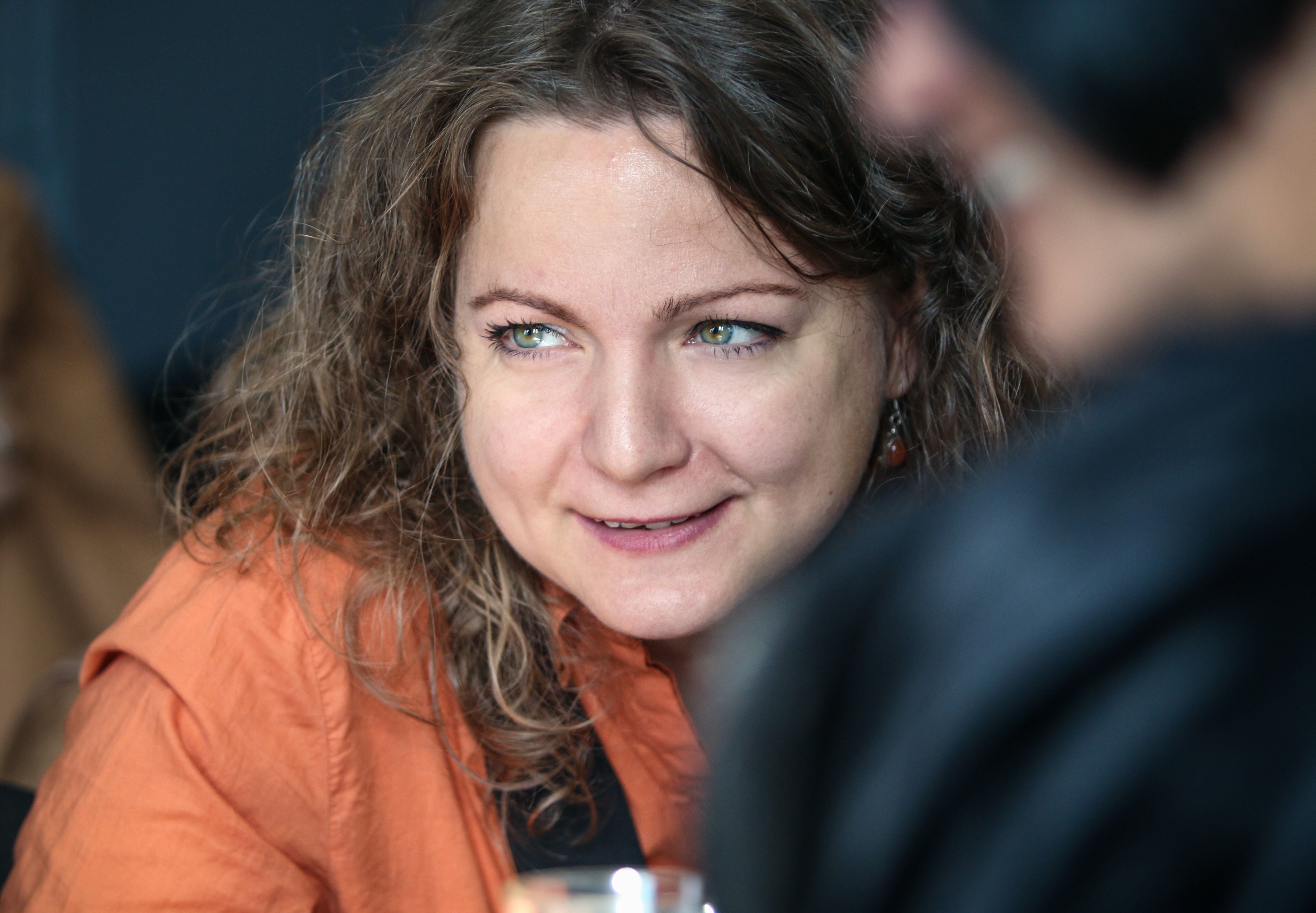 Eine Frau mit lockigem Haar und orangefarbener Bluse lächelt und blickt zur Seite.