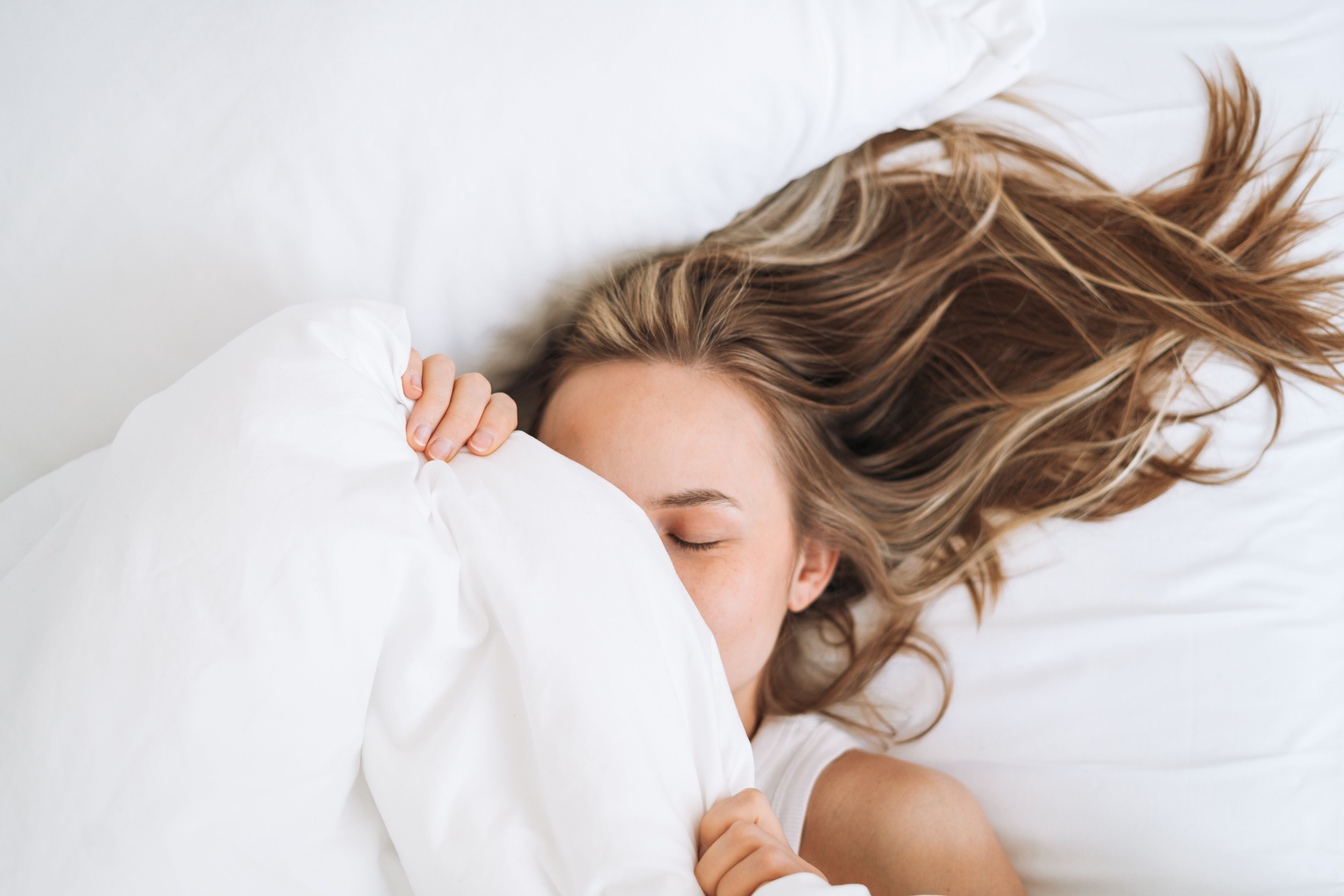 Junge Frau mit langen Haaren liegt entspannt mit geschlossenen Augen im weißen Bett.