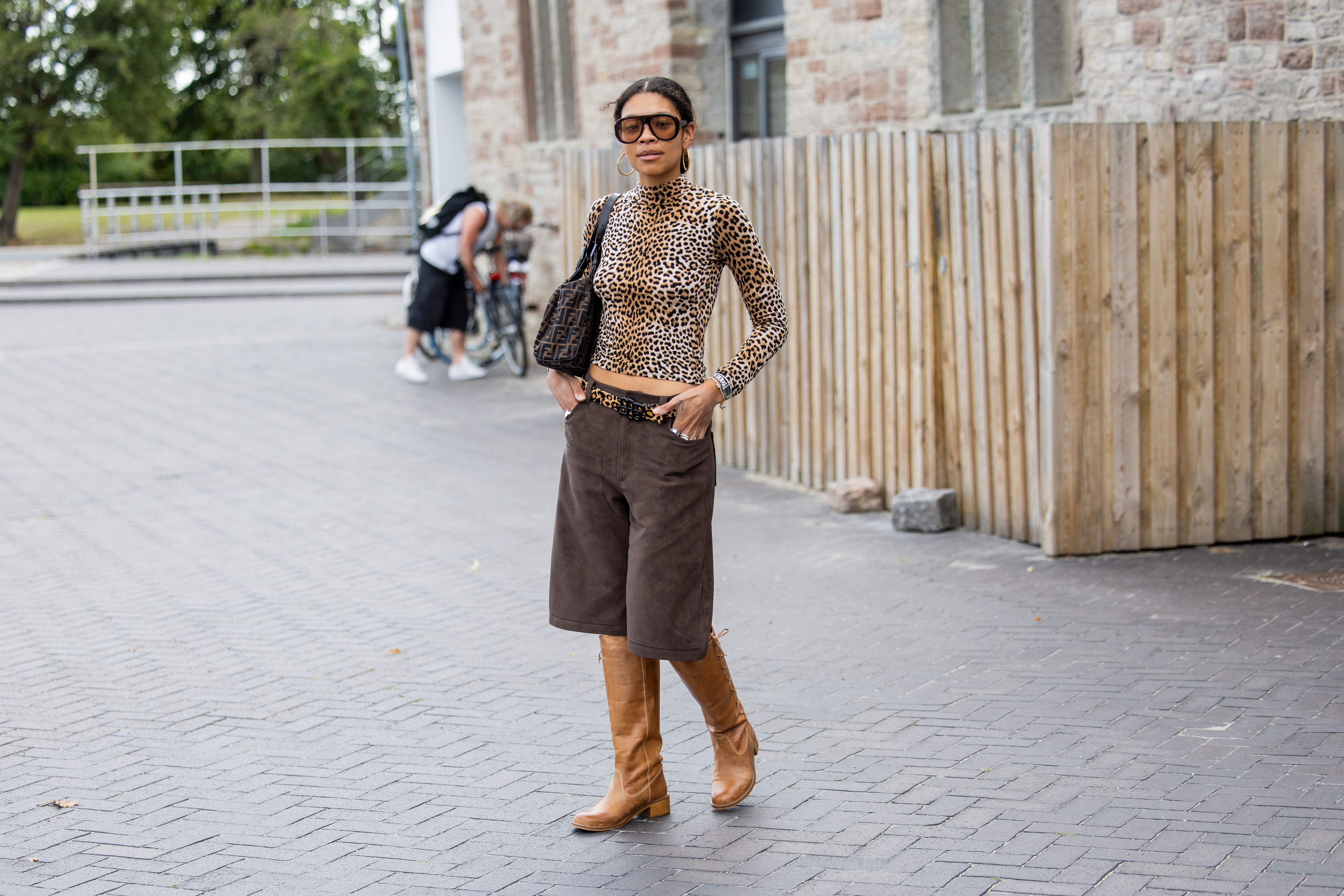 Frau mit Animalprint-Top, braunen Shorts und Stiefeln posiert modisch auf der Straße.