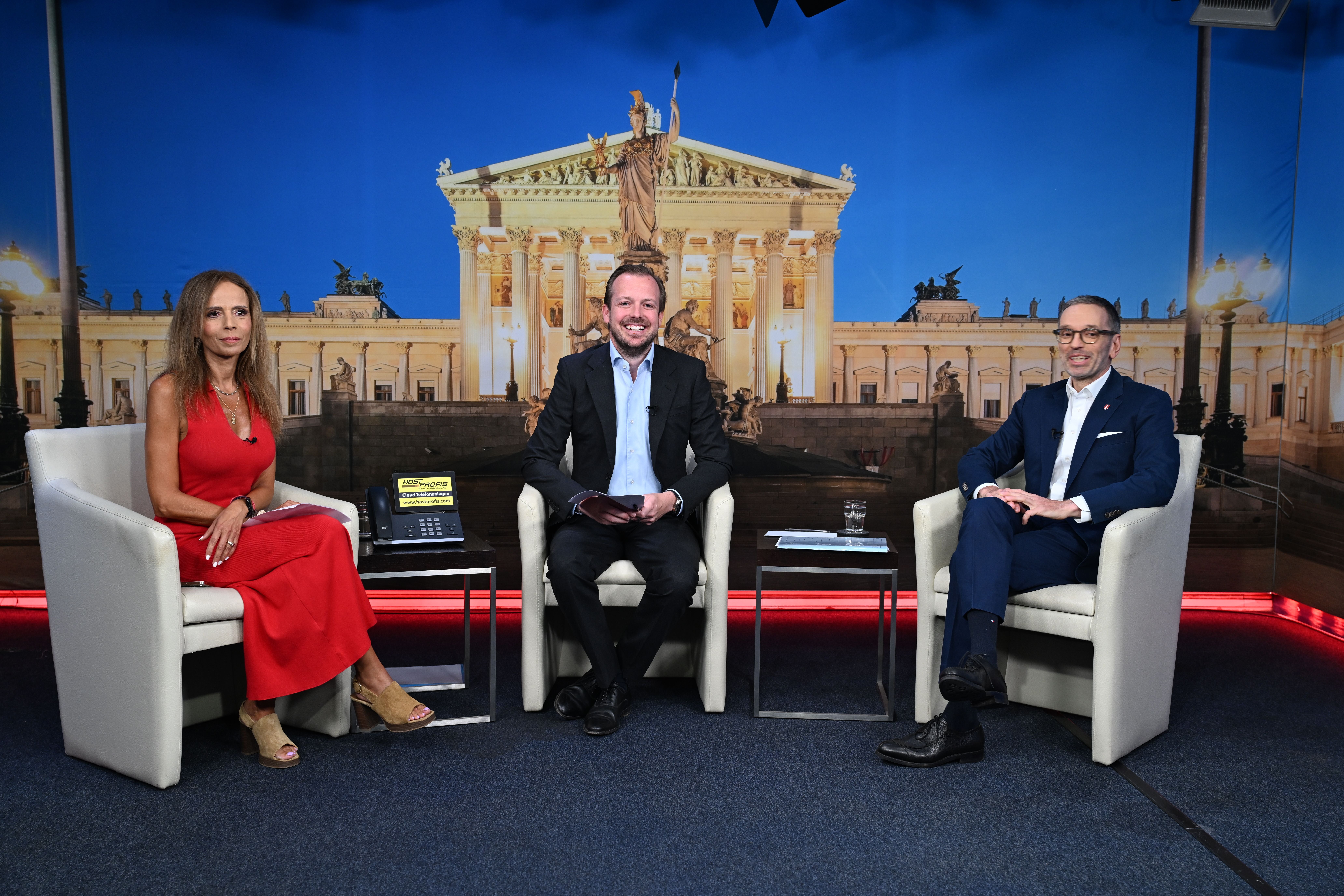 Drei Personen sitzen in einem Fernsehstudio vor einem Hintergrundbild des Parlamentsgebäudes.