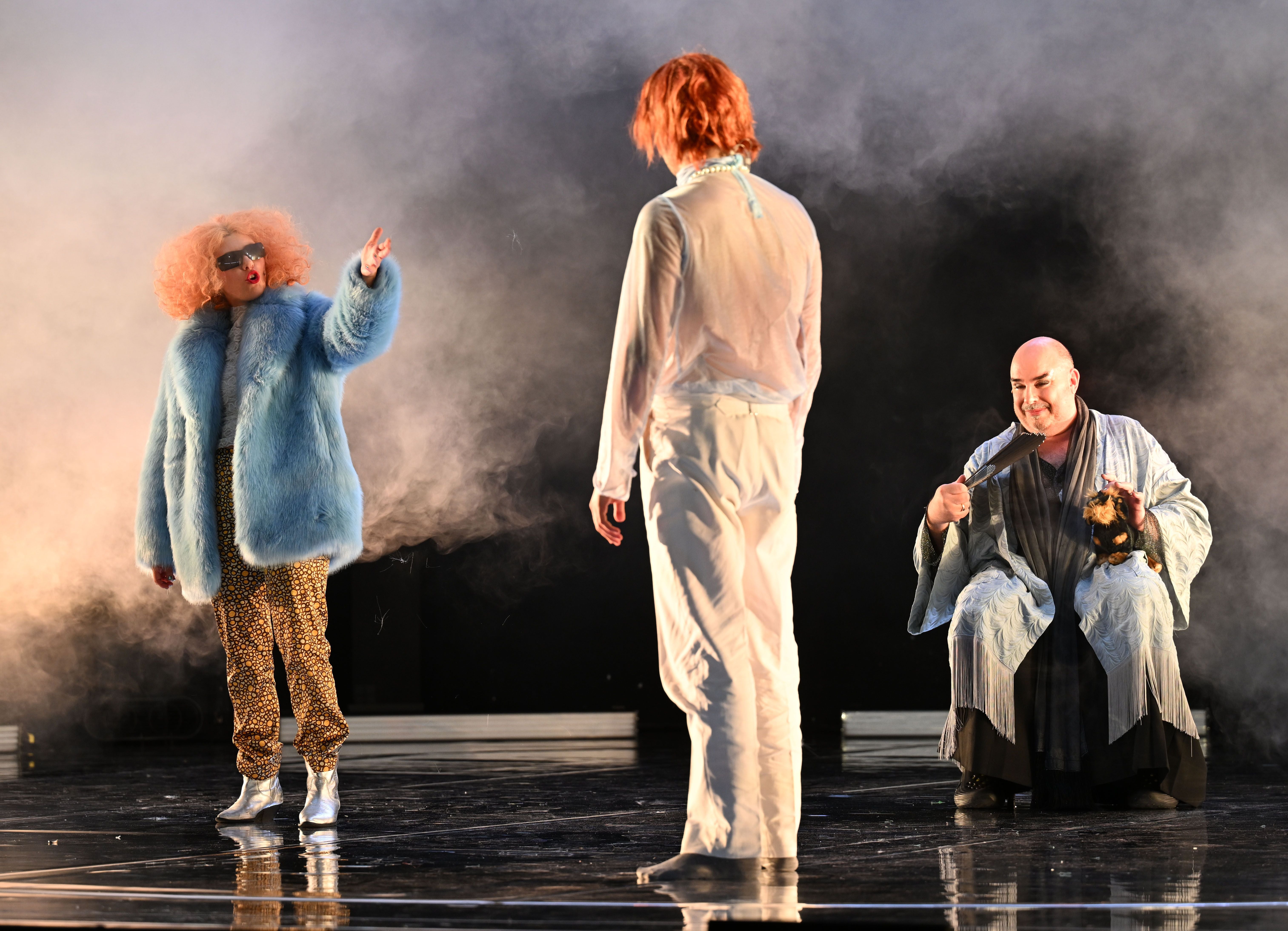 Drei Schauspieler:innen auf Theaterbühne in bunten Kostümen, Bühnennebel im Hintergrund.