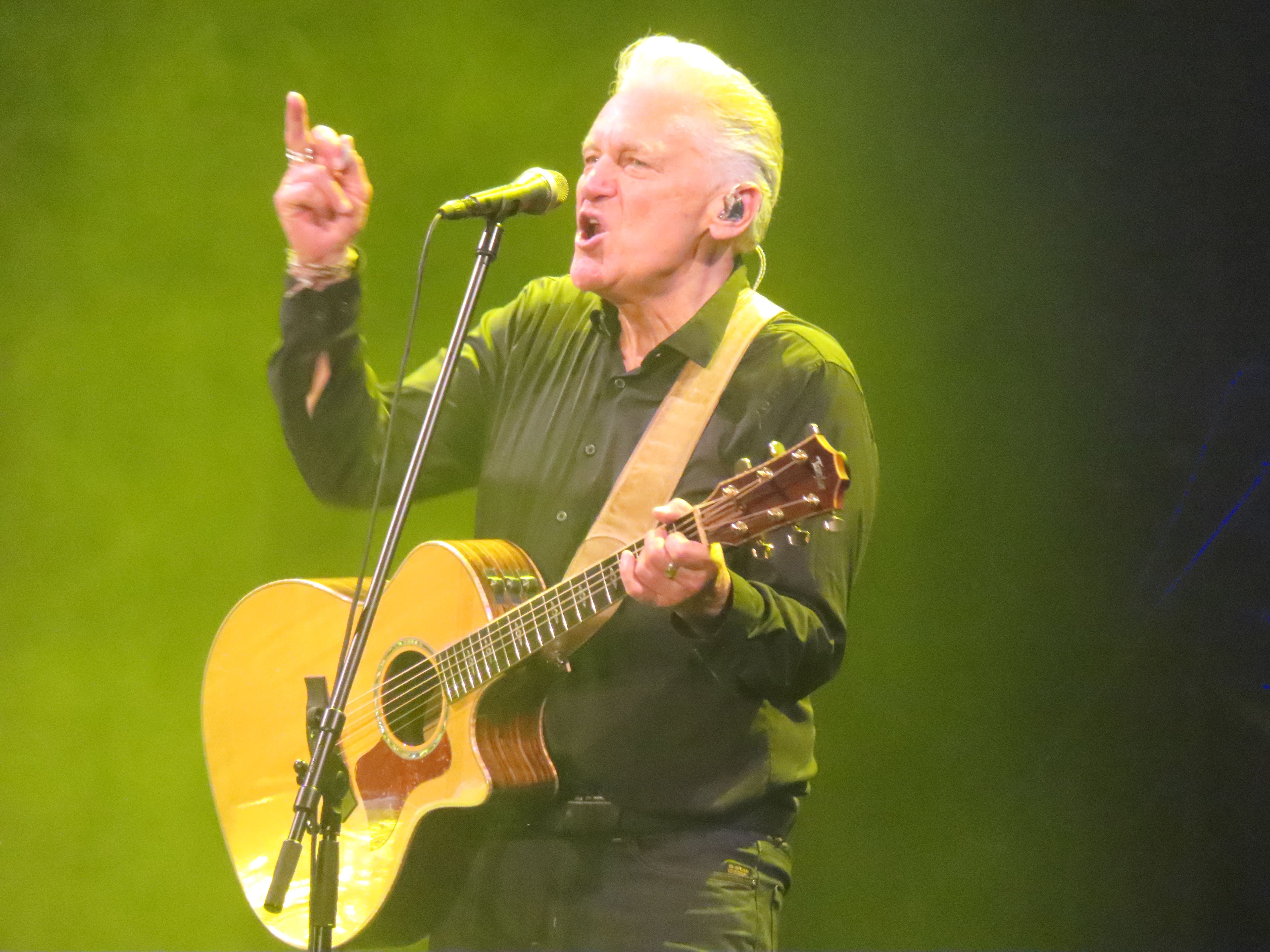 Ein älterer Mann spielt Gitarre und singt auf einer Bühne im gelben Licht.