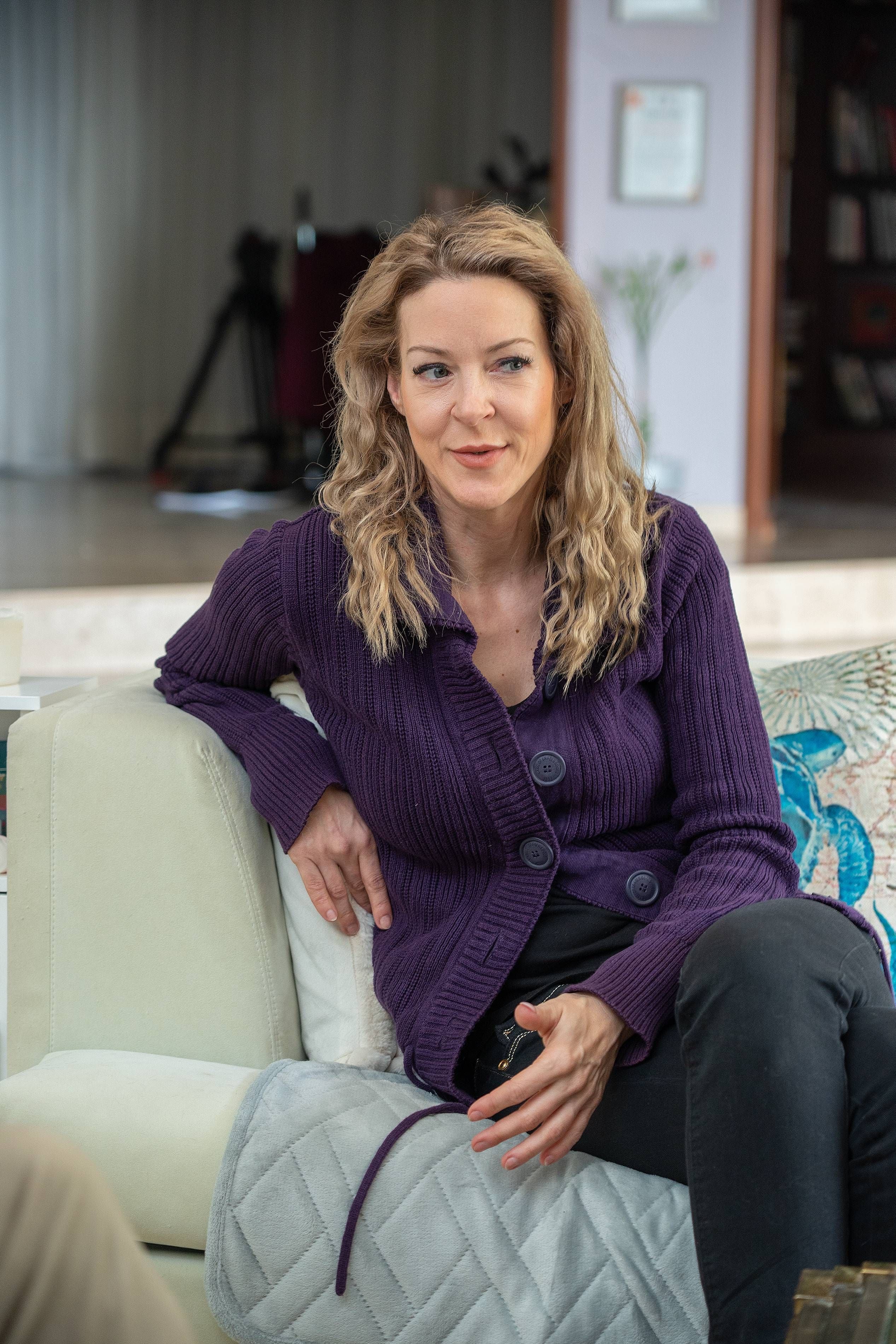 Frau mit langen blonden Haaren sitzt auf einem Sofa und trägt einen lila Strickpullover.
