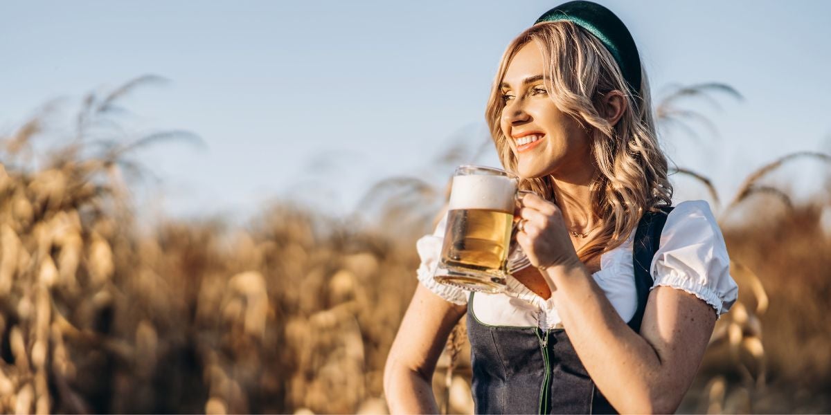 Frau in Dirndl hält Bierkrug und lächelt im Sonnenschein auf einem Feld.