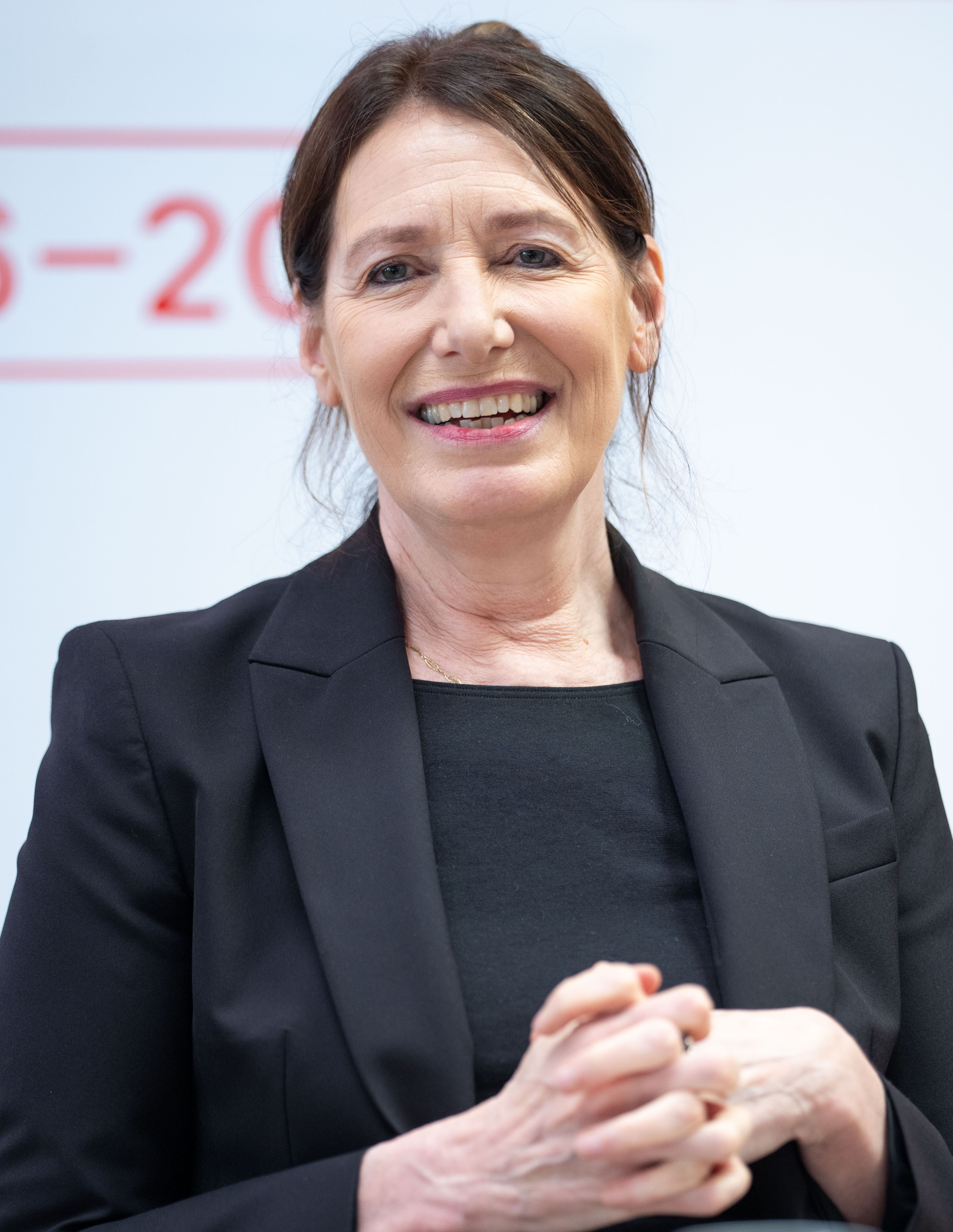 Eine lächelnde Frau im schwarzen Blazer bei einer Pressekonferenz in Wien.