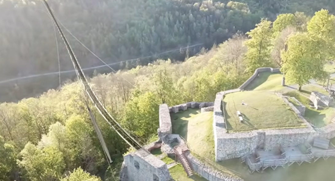 Hängebrücke neben einer alten Burgruine im bewaldeten Tal, aus der Vogelperspektive fotografiert.