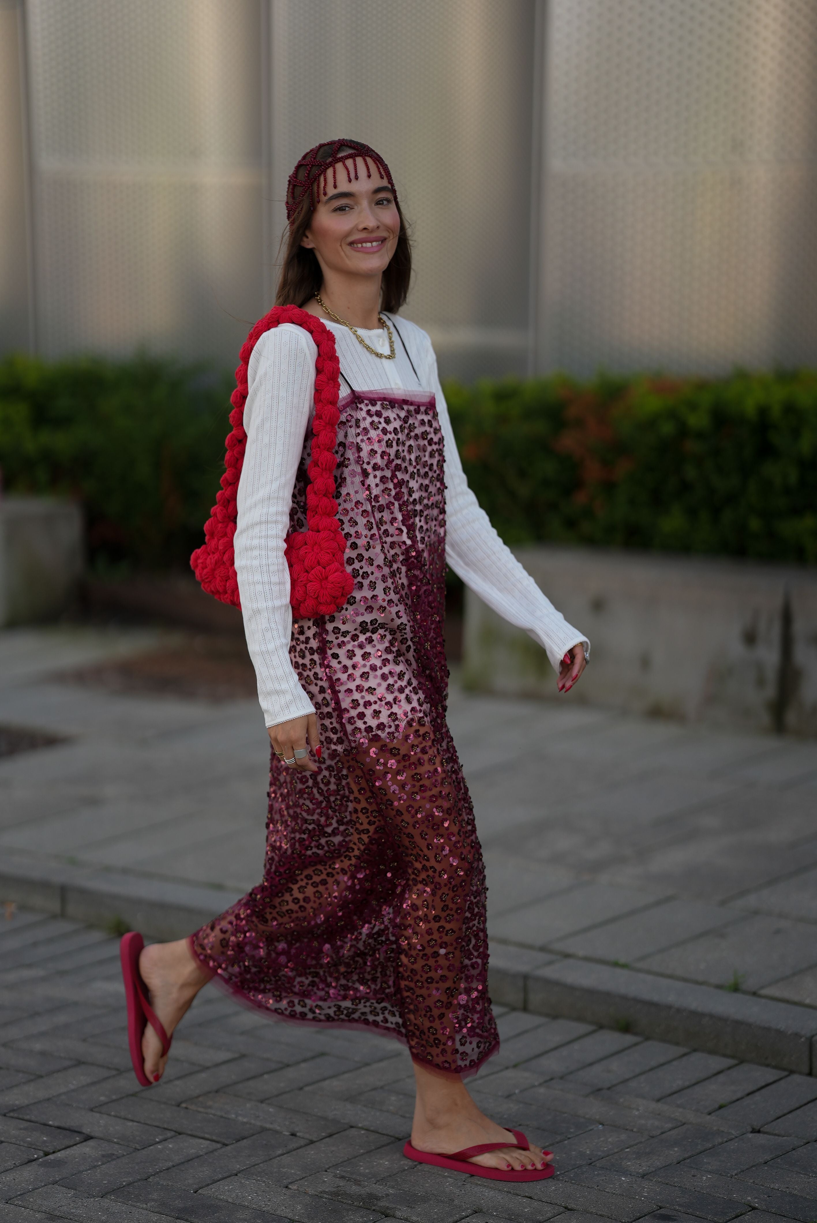 Frau mit burgunderfarbenem Paillettenkleid, roter Stricktasche und Flip-Flops auf der Straße.