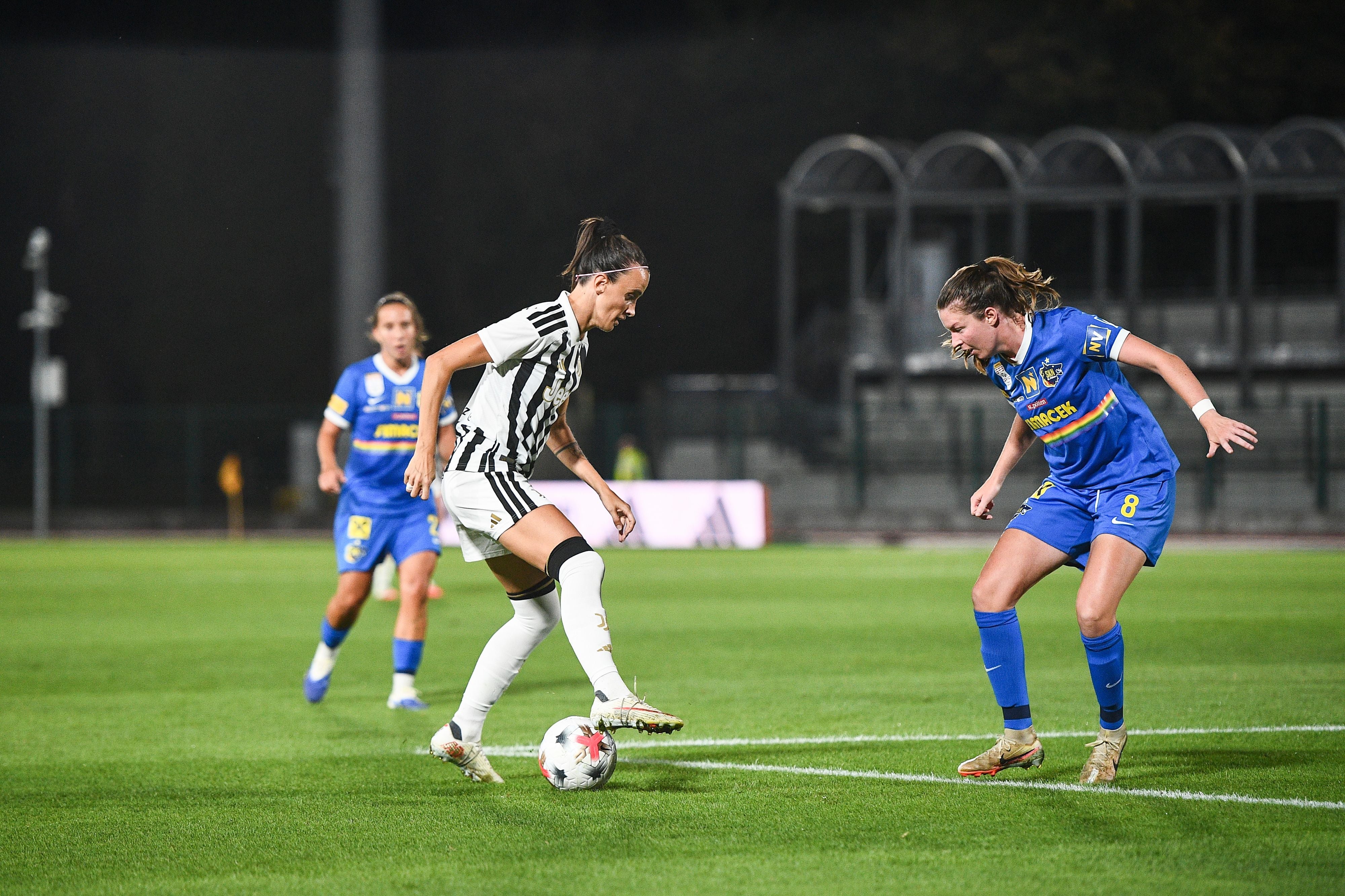 Eine Juventus-Spielerin dribbelt den Ball am Fuß und wird dabei von einer St. Pölten-Spielerin im blauen Trikot attackiert. Eine weitere Spielerin beobachtet die Szene im Hintergrund auf dem Rasenplatz bei Flutlicht.