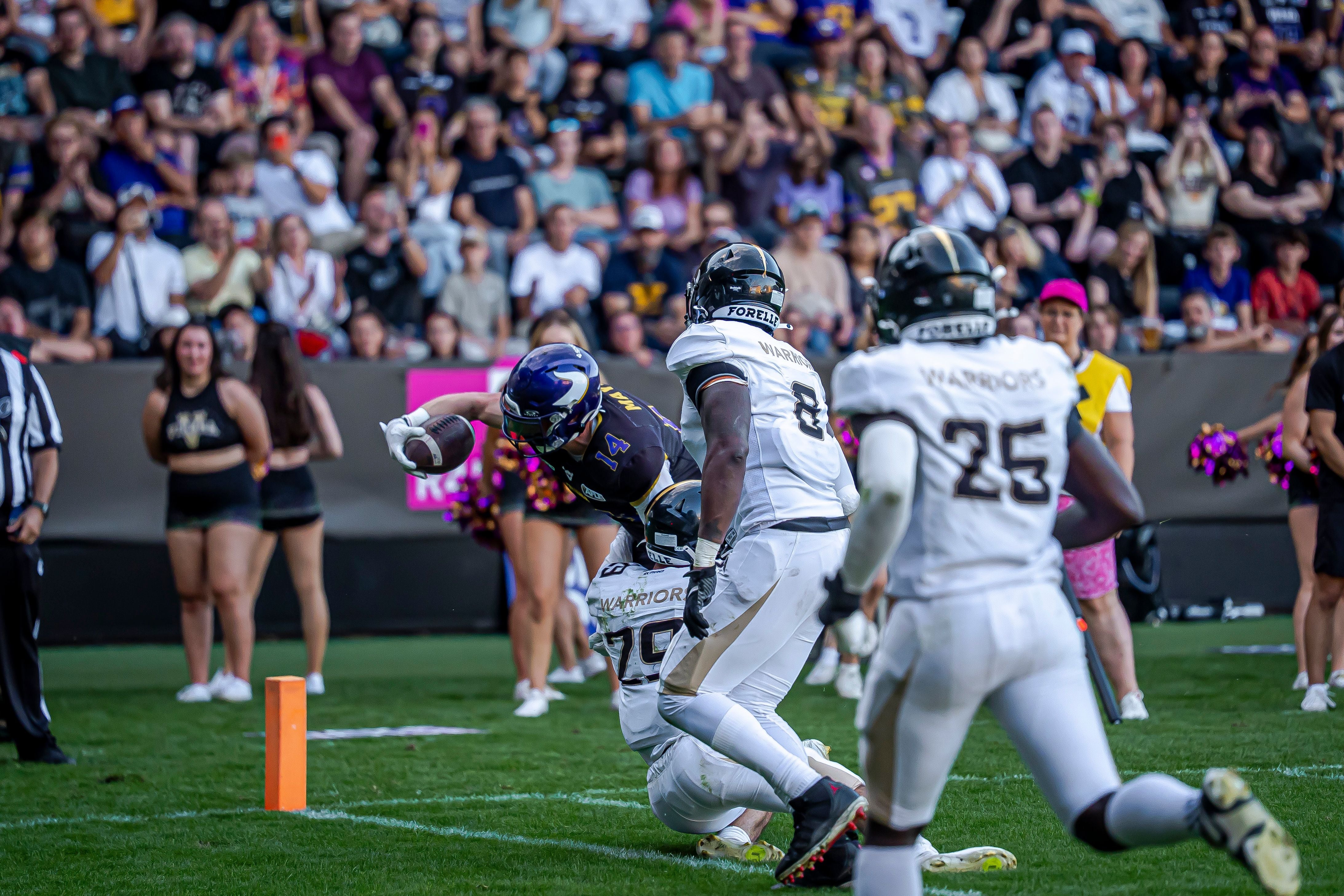 Ein Footballspieler streckt den Ball Richtung Endzone, während er von Gegnern getackelt wird.