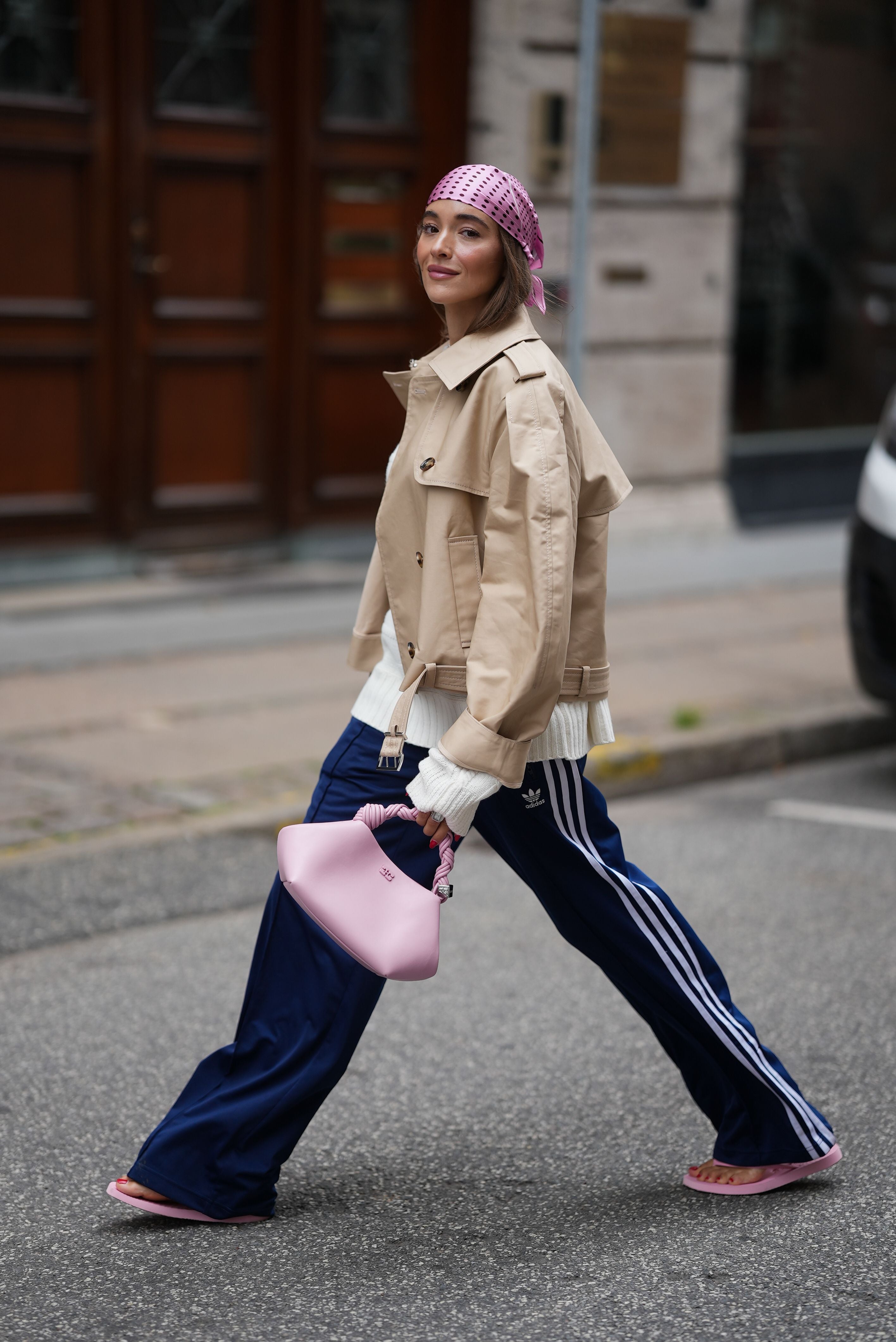Frau mit beigem Trenchcoat, blauer Sporthose, rosa Kopftuch und pinker Tasche auf der Straße.