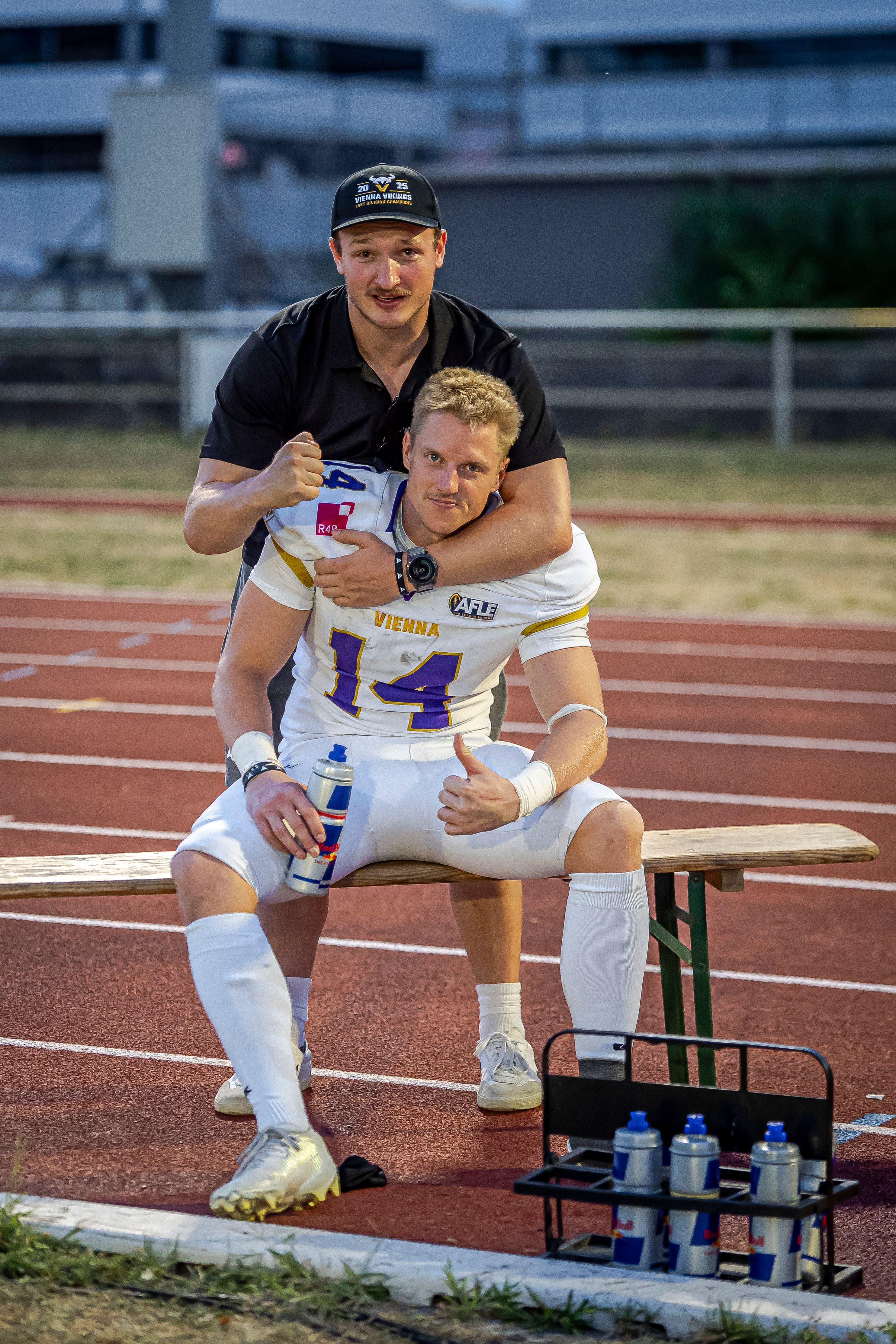 Zwei Männer, einer im Football-Outfit, sitzen und posieren auf einer Bank am Sportplatz.