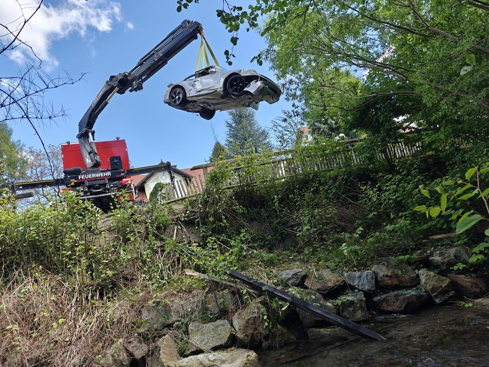 Ein Feuerwehrkran hebt ein verunfalltes Auto aus einem bewaldeten Bachbett.