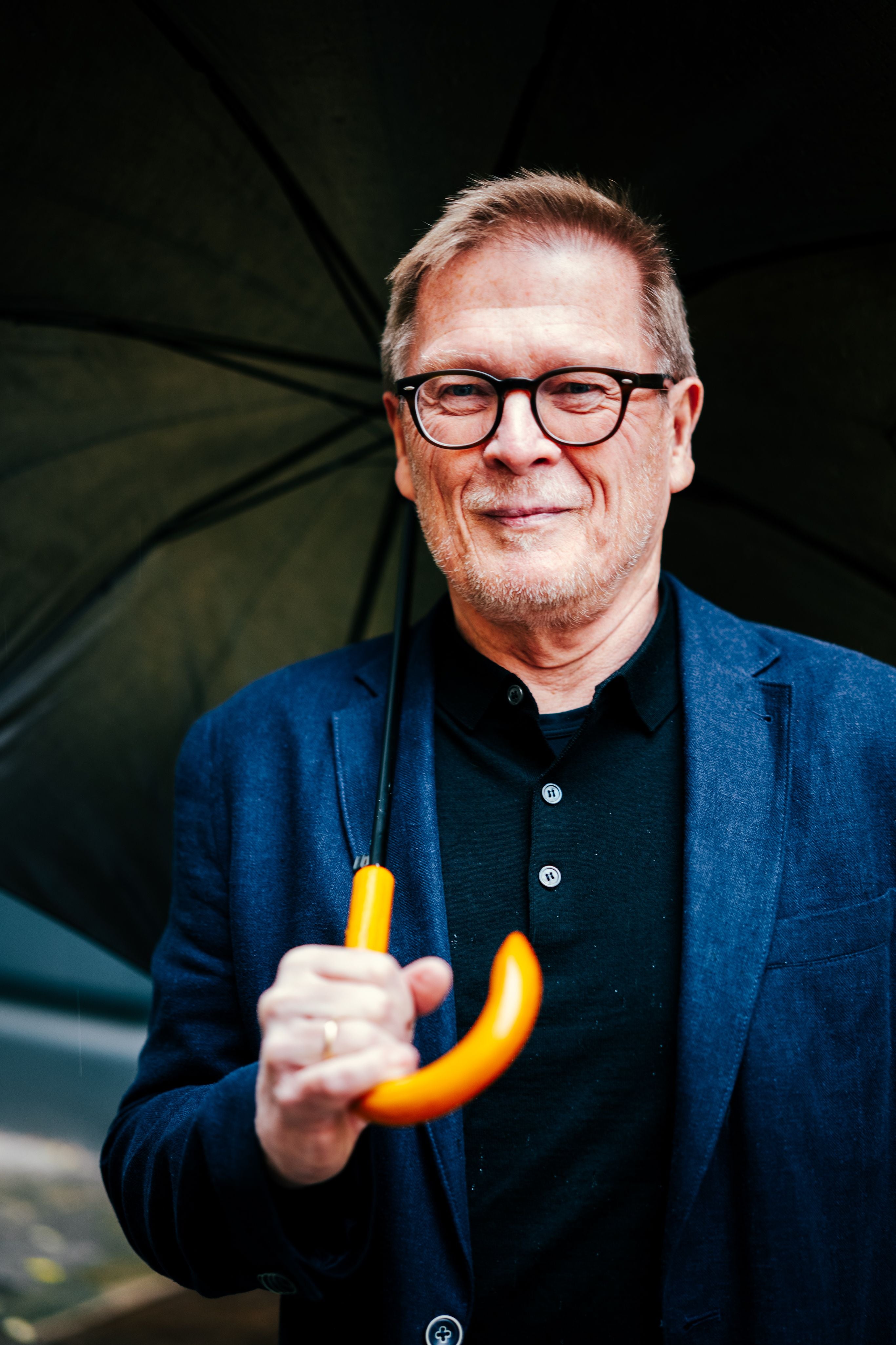 Ein älterer Mann mit Brille hält einen schwarzen Regenschirm mit orangem Griff.