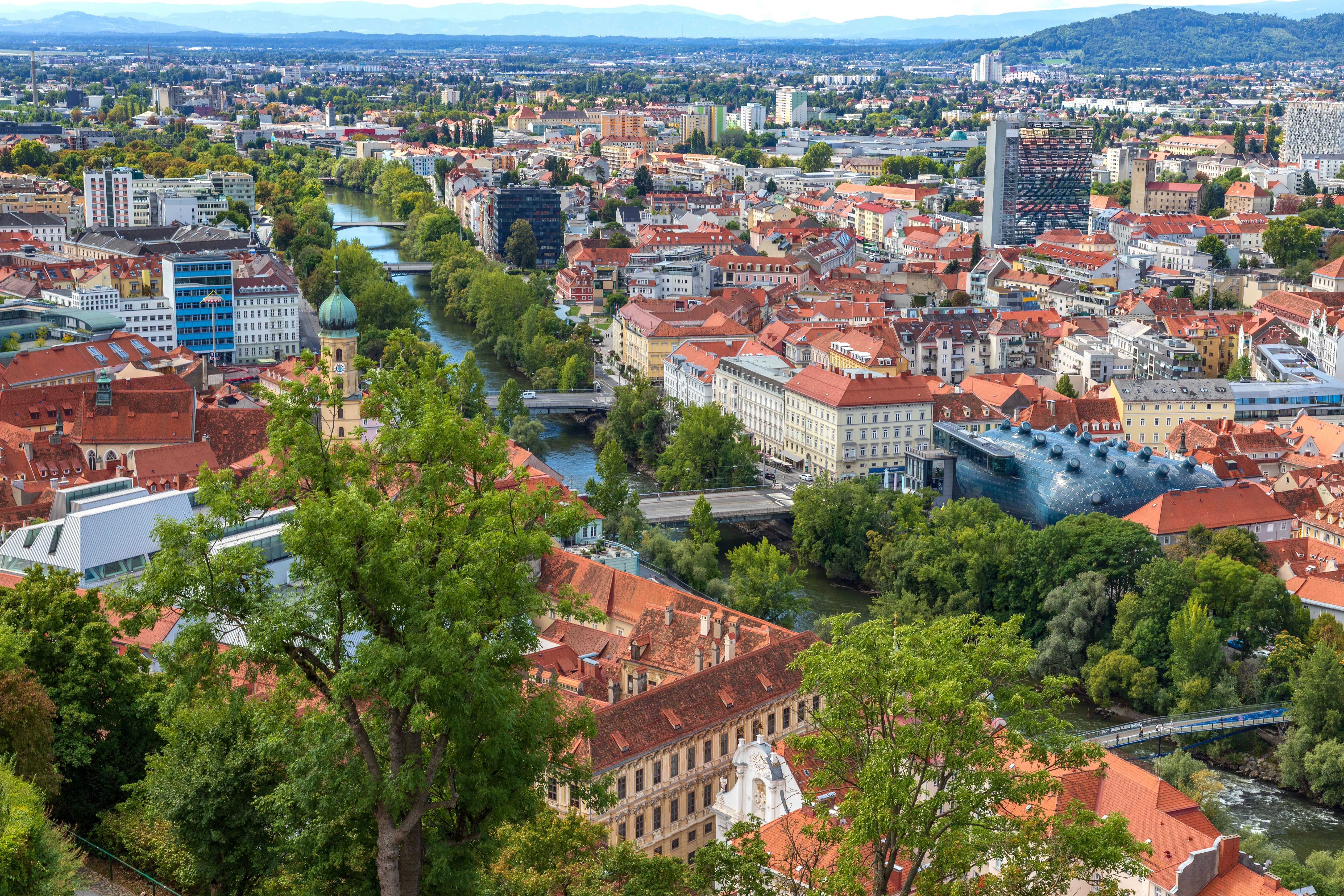 Blick auf Graz mit rotem Ziegeldächern, der Mur und dem modernen Kunsthaus am Fluss.