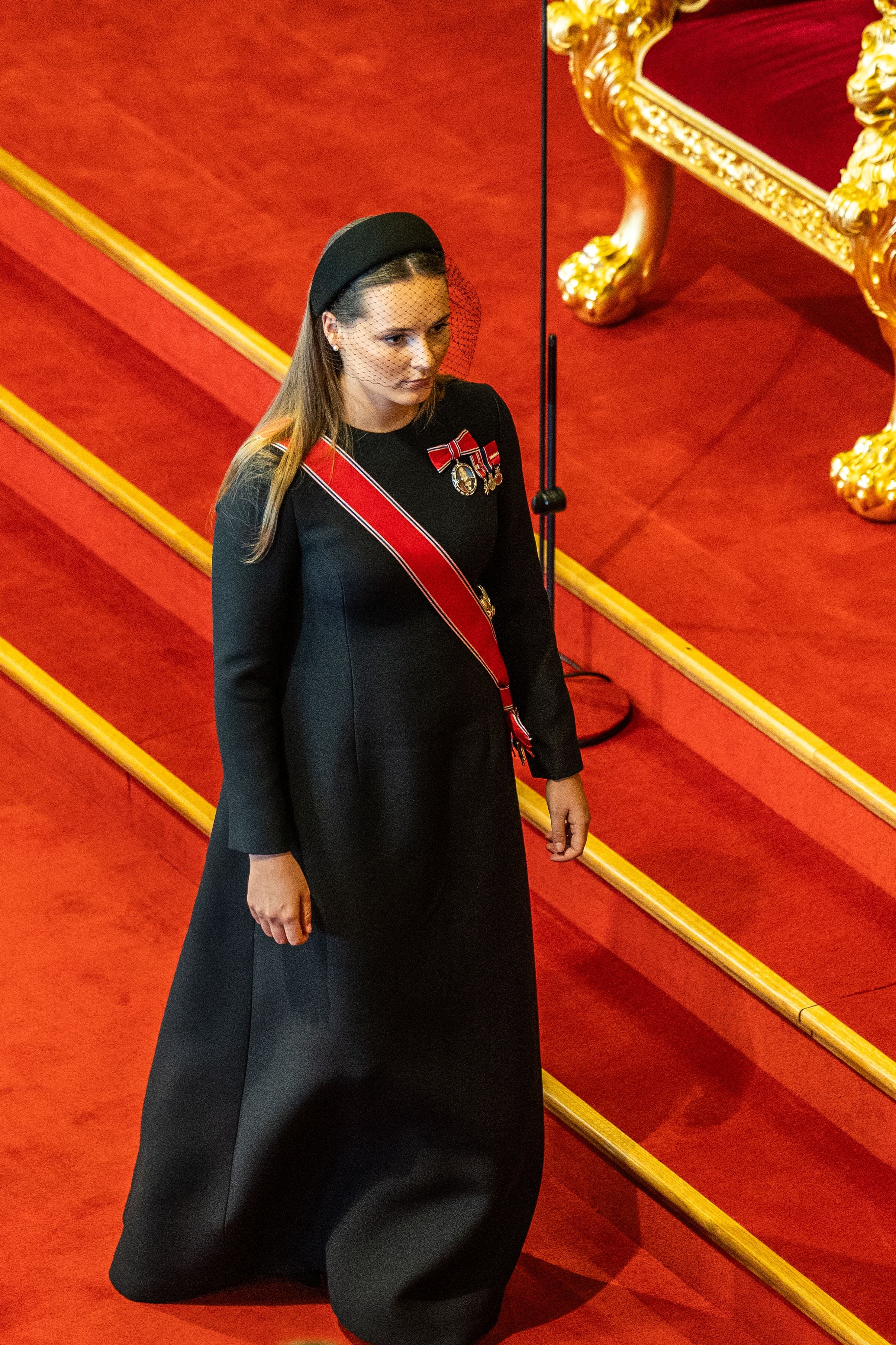 Eine Frau im schwarzen Kleid mit Schärpe und Orden steht auf rotem Teppich in einem prunkvollen Saal.