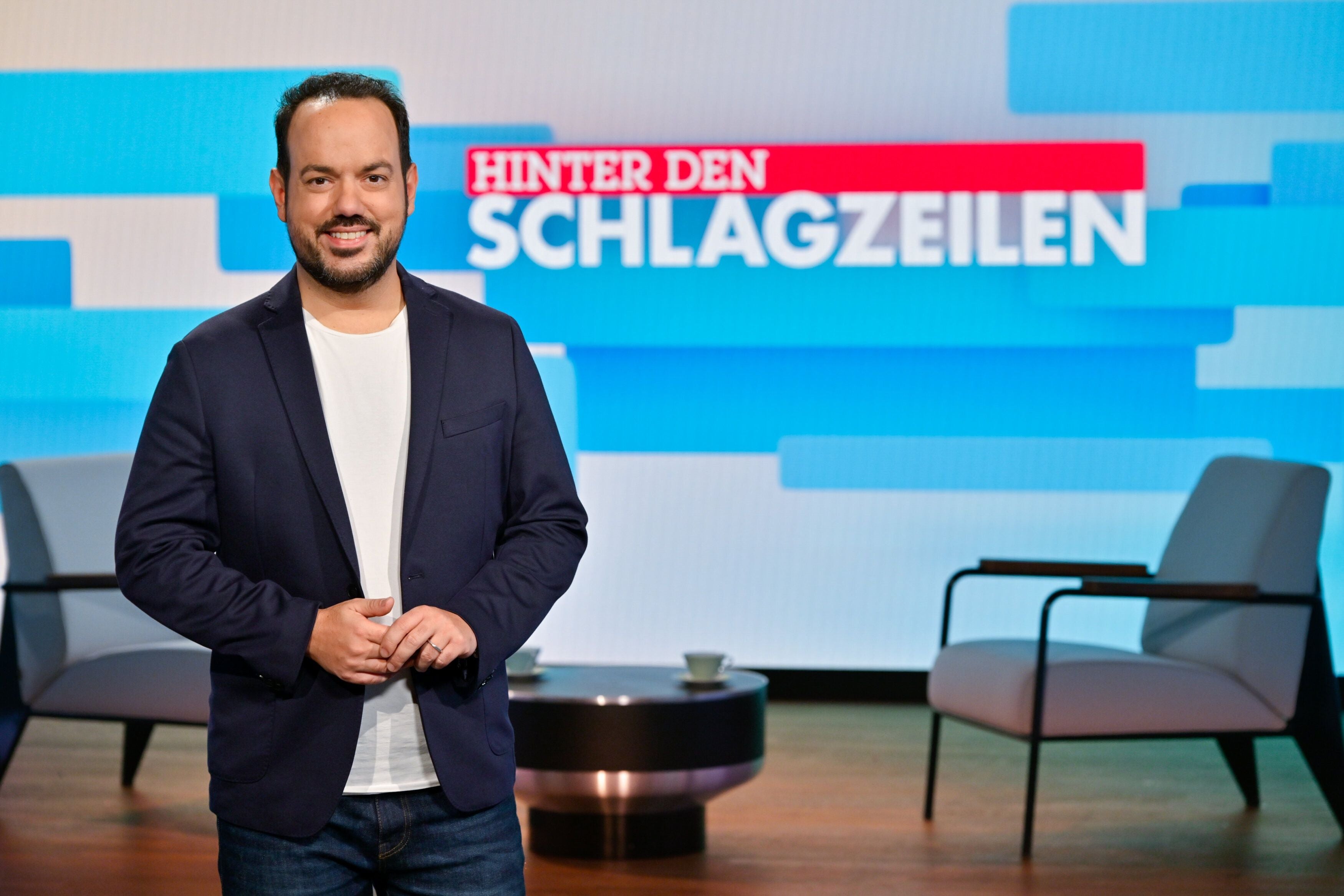 Ein Mann im Studio steht vor der Tafel mit dem Schriftzug „Hinter den Schlagzeilen“.