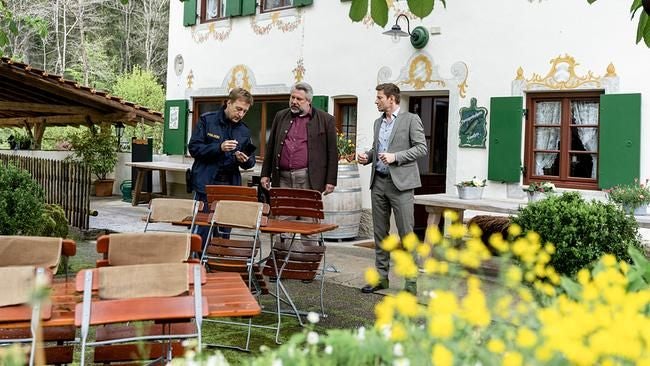 Drei Männer unterhalten sich im Garten eines traditionellen Hauses mit grünen Fensterläden.