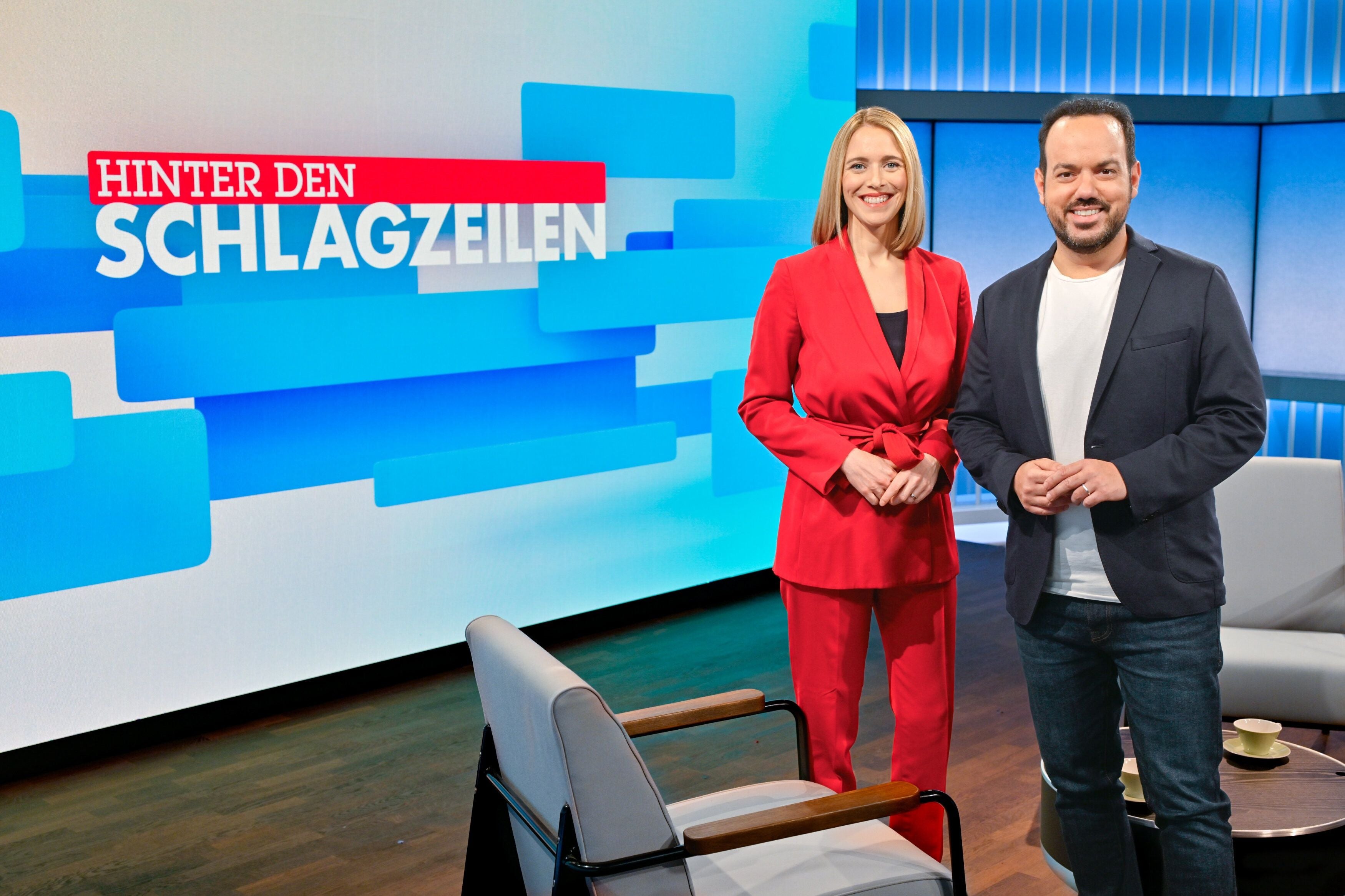 Zwei Moderatoren stehen im TV-Studio vor dem „Hinter den Schlagzeilen“-Sendungslogo.