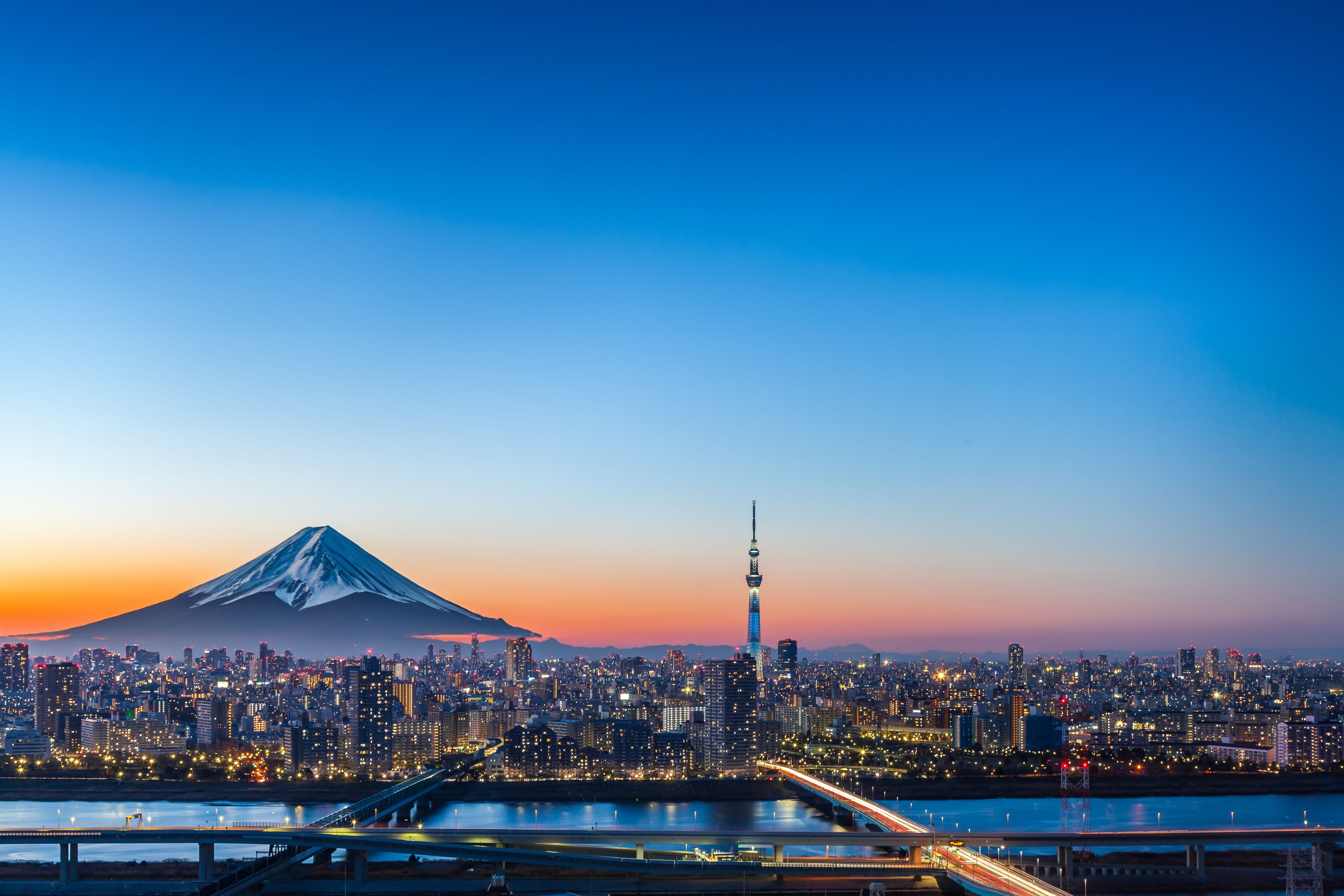 Stadtansicht von Tokio bei Sonnenuntergang mit Mount Fuji im Hintergrund und beleuchteten Gebäuden.