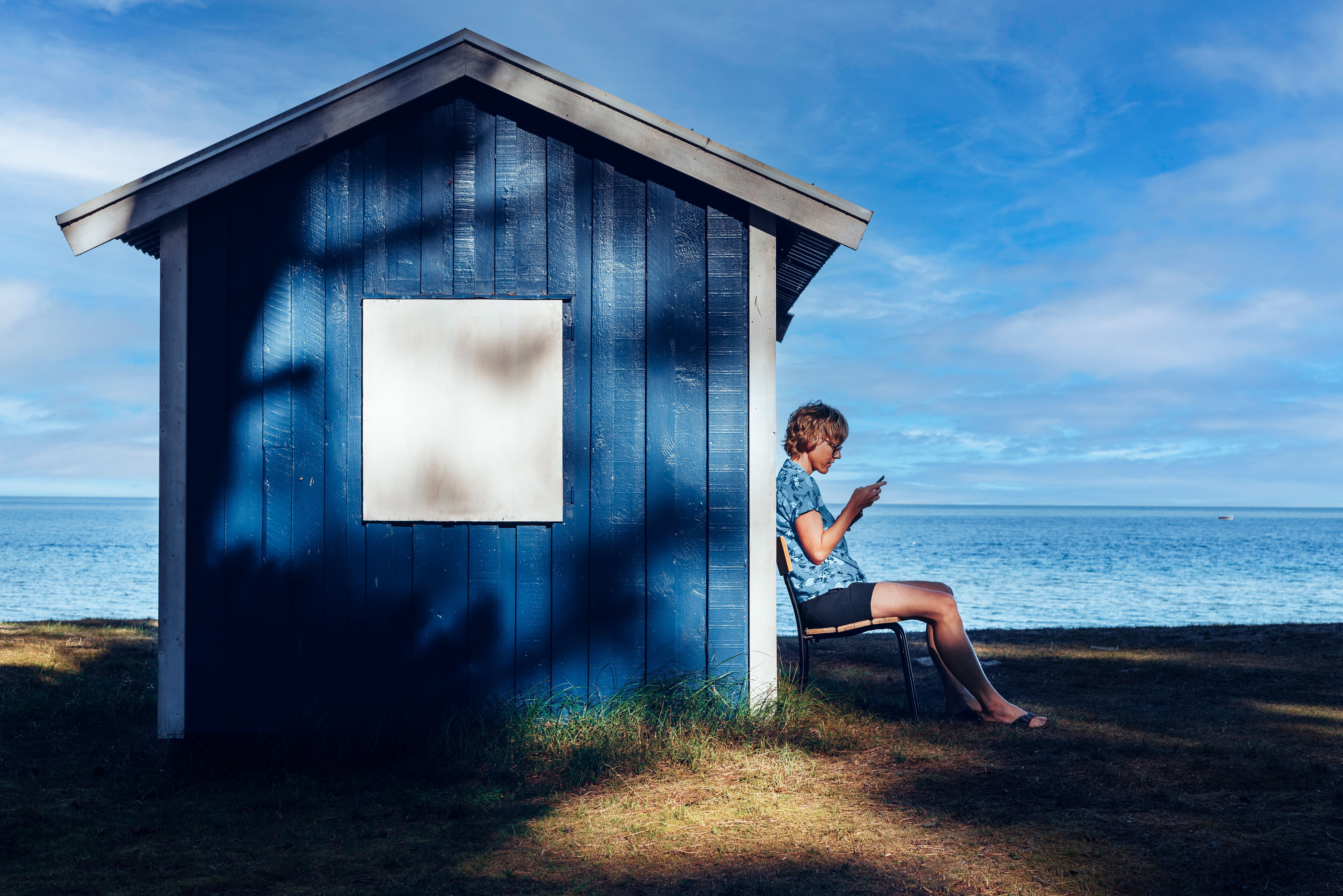 Person sitzt auf einem Stuhl neben einer blauen Hütte und schaut auf ein Handy am Meer.