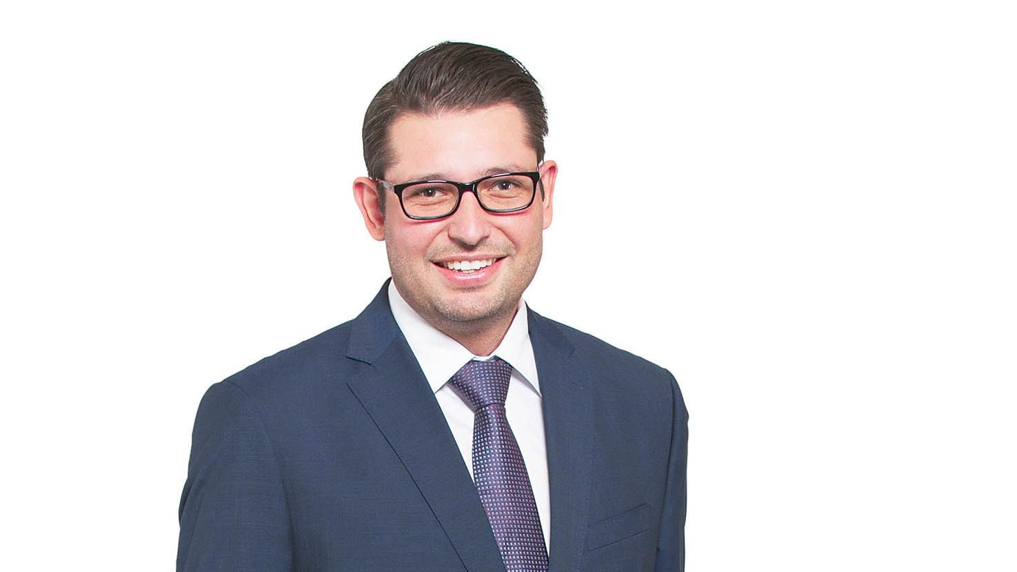 Mann im Anzug und mit Brille lächelt vor weißem Hintergrund in die Kamera.