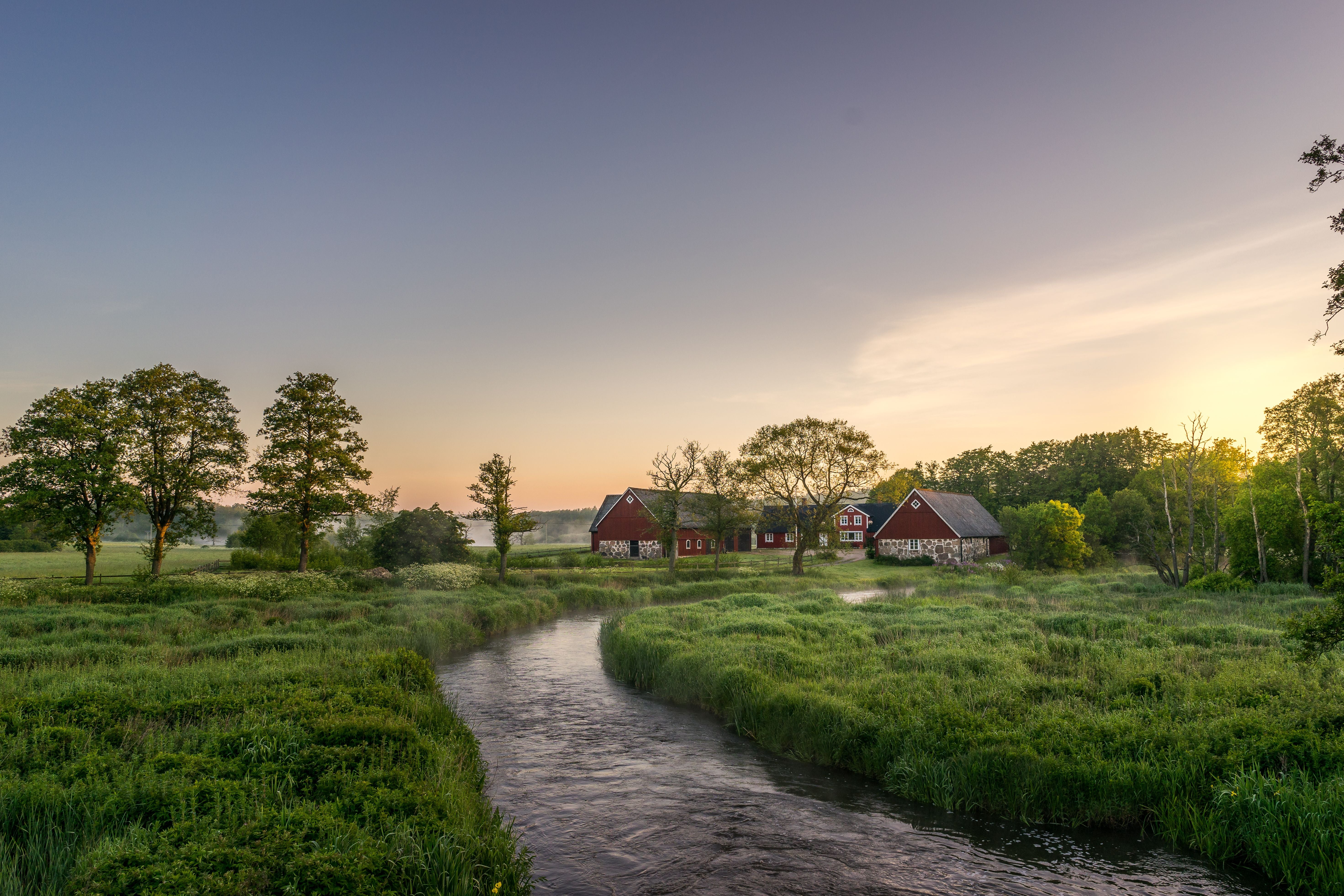 Idyllischer Bauernhof mit roten Gebäuden und Fluss in südschwedischer Landschaft bei Sonnenuntergang.