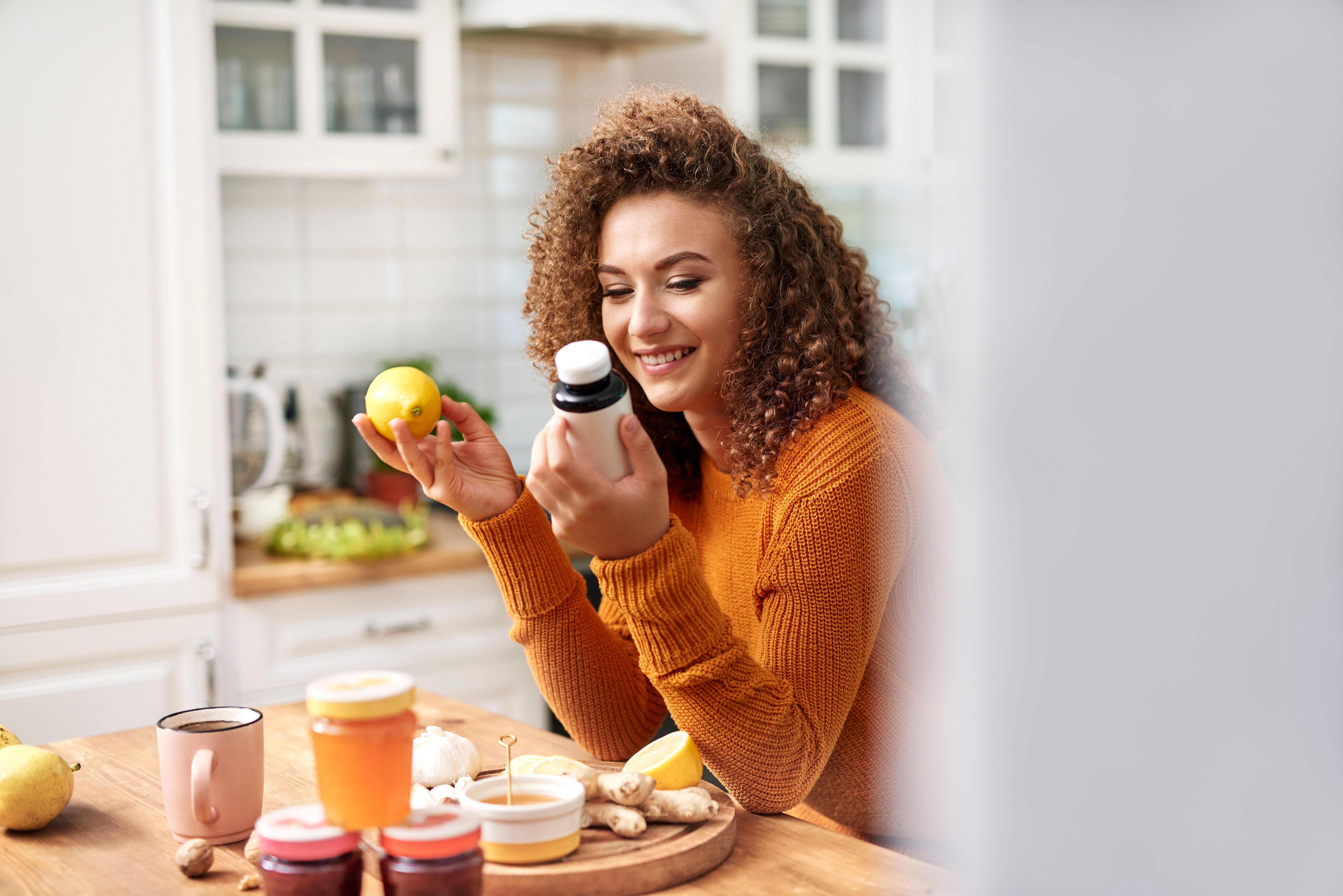 Junge Frau sitzt in der Küche und vergleicht eine Zitrone mit einer Vitaminflasche.