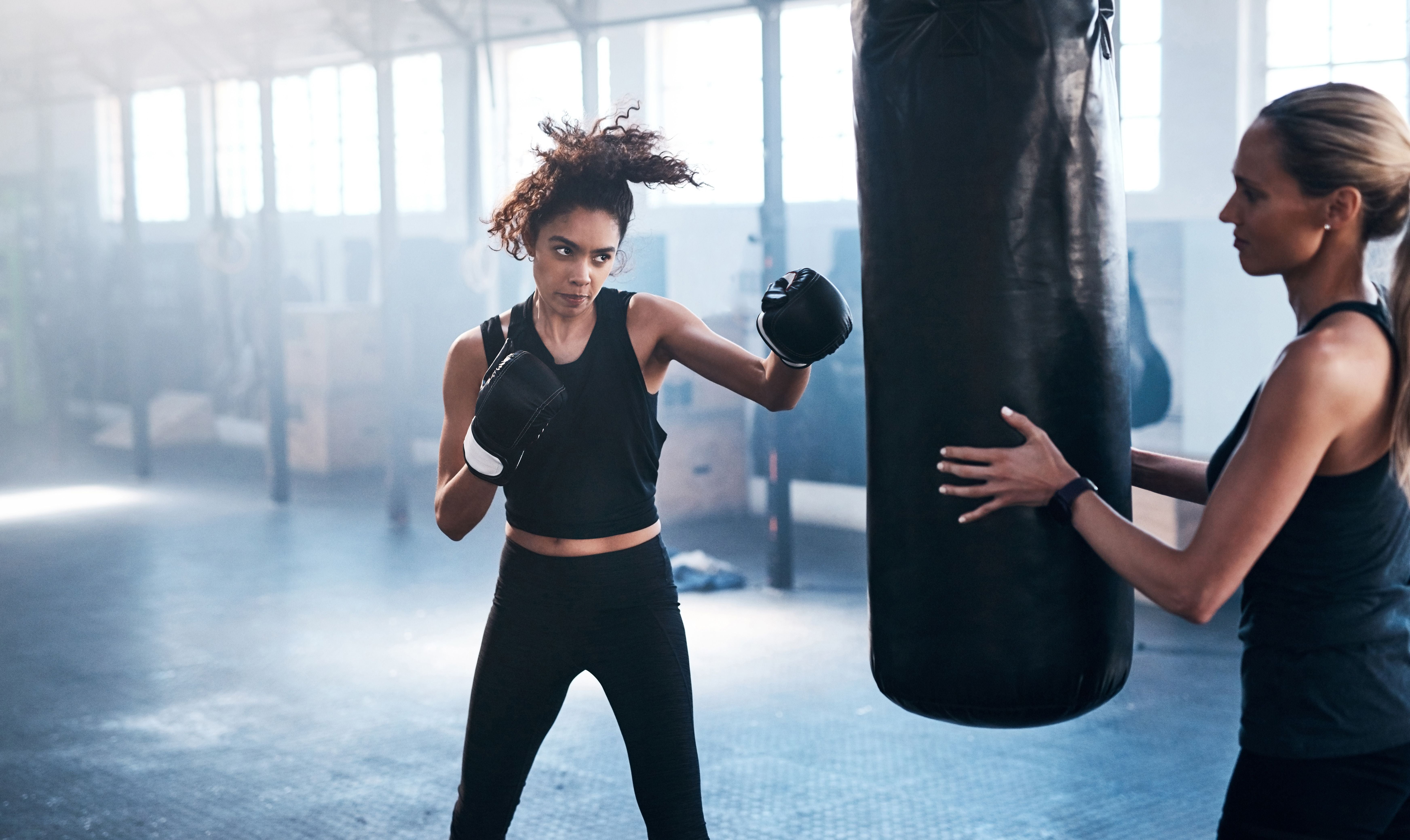 Zwei Frauen trainieren Boxen im Fitnessstudio, eine schlägt auf einen Sandsack, die andere hält ihn fest.