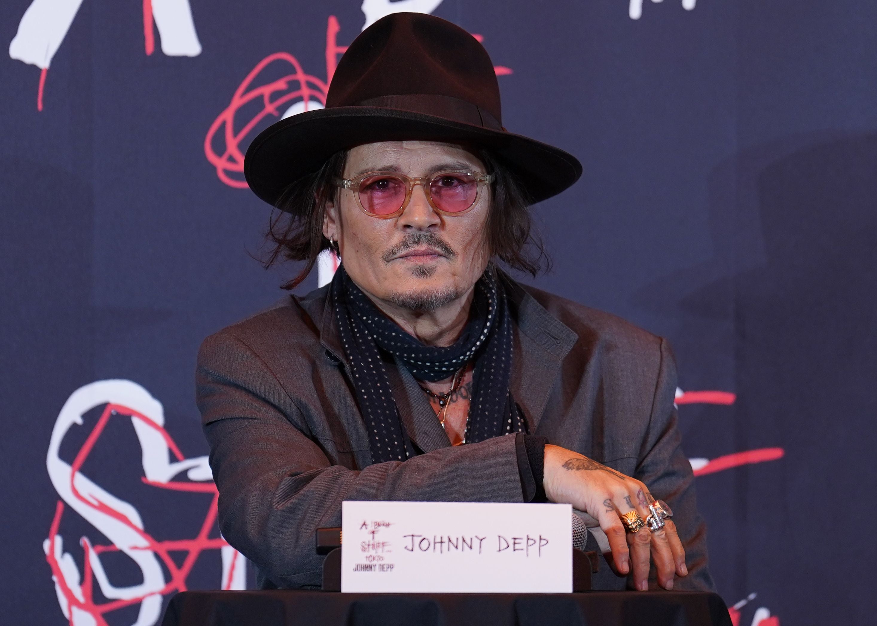 Mann mit Hut, Sonnenbrille und Schal sitzt bei einer Pressekonferenz an einem Tisch mit Namensschild.