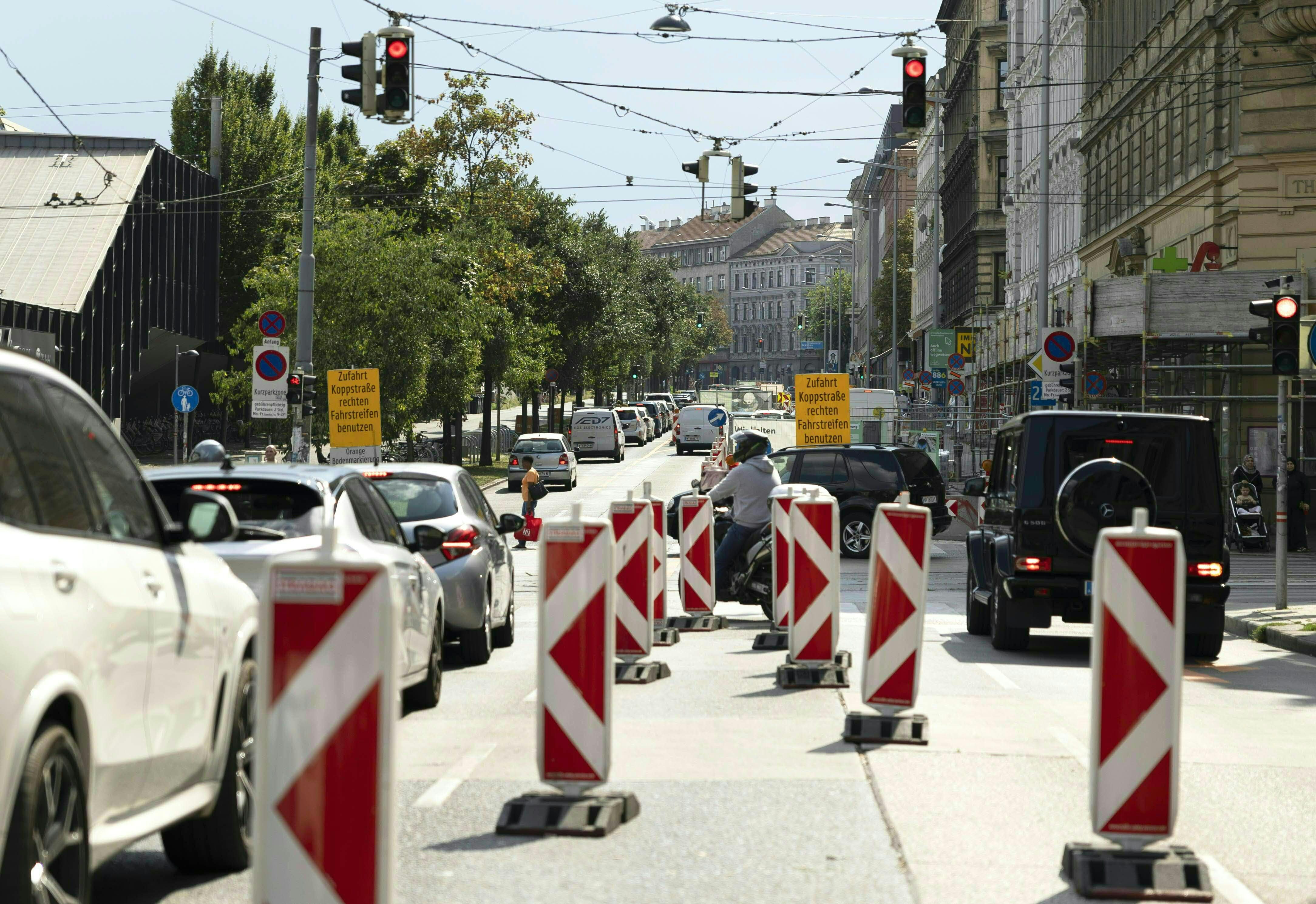Autos und Motorräder stehen vor einer Baustelle mit Absperrungen und roten Ampeln in einer Stadtstraße.