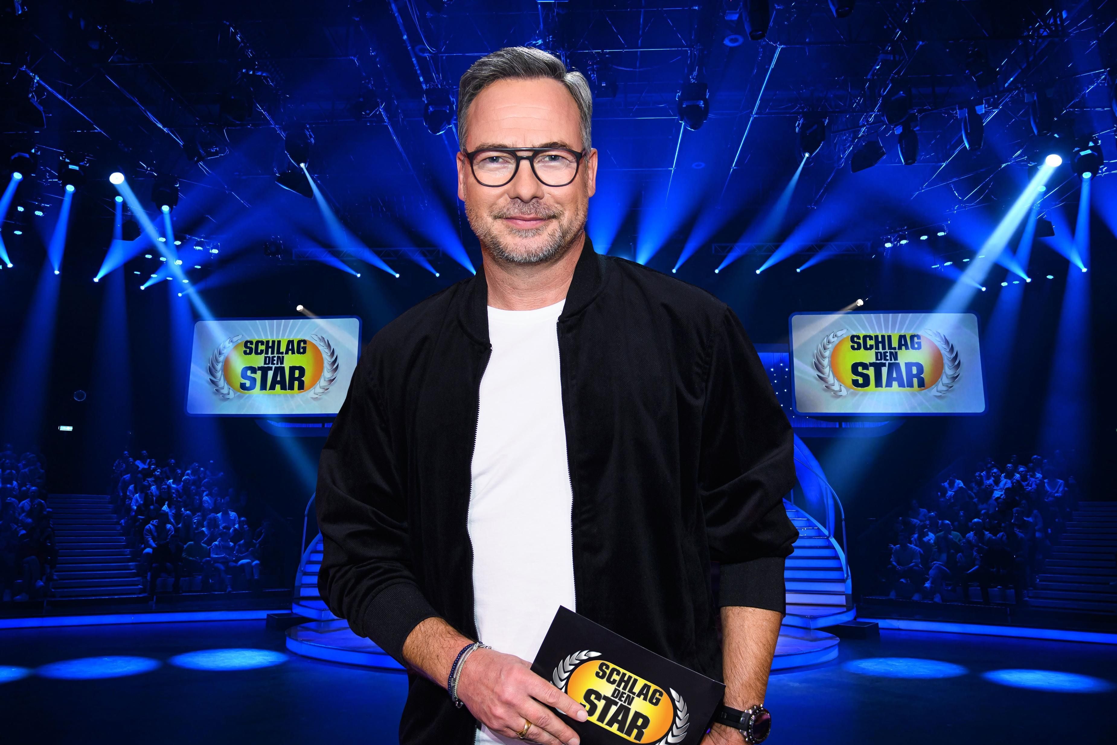 Ein Mann mit Brille steht auf der Bühne von "Schlag den Star" mit Publikum im Hintergrund.