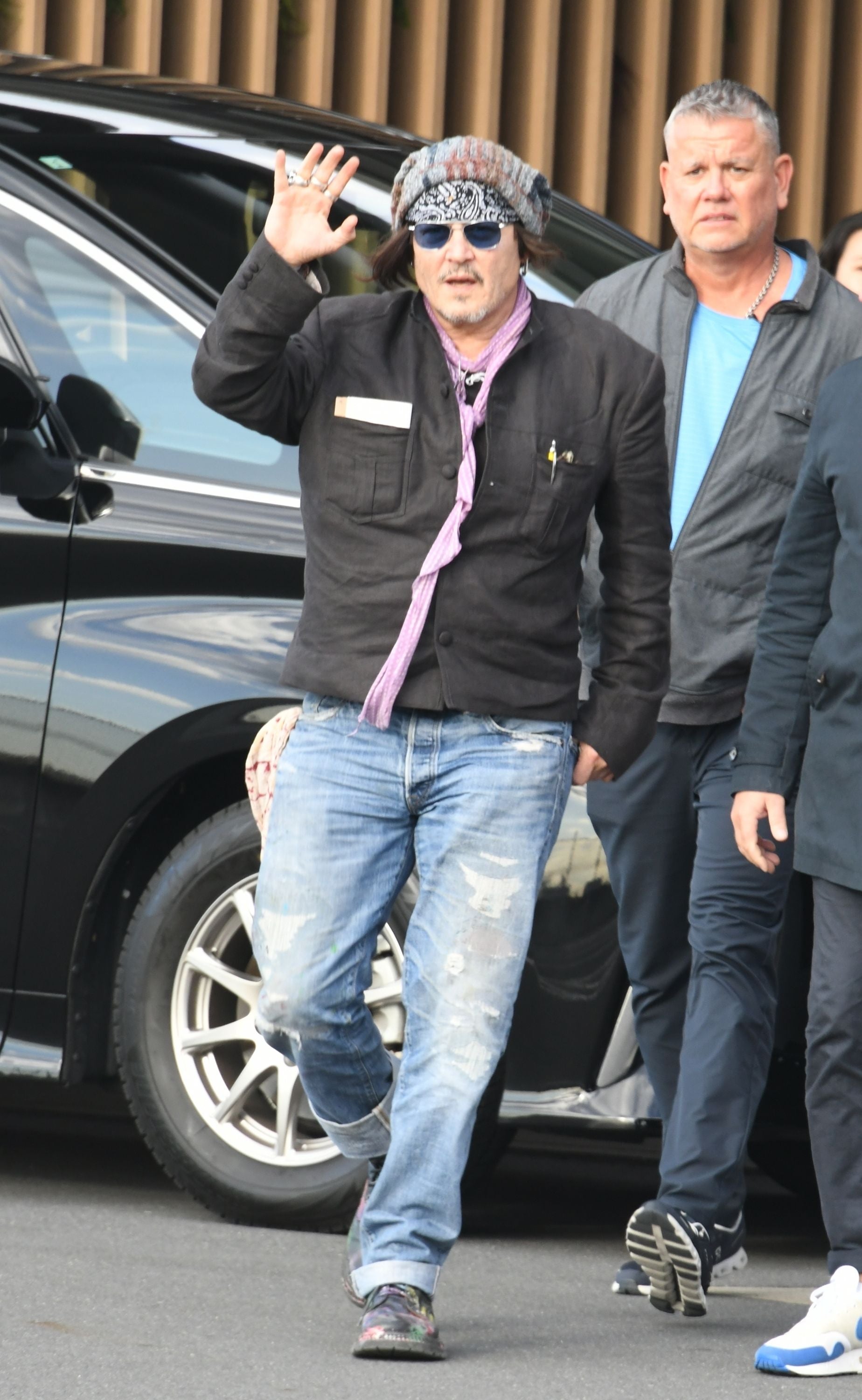Ein Mann mit Sonnenbrille und Mütze winkt beim Aussteigen aus einem Auto, weitere Personen im Hintergrund.