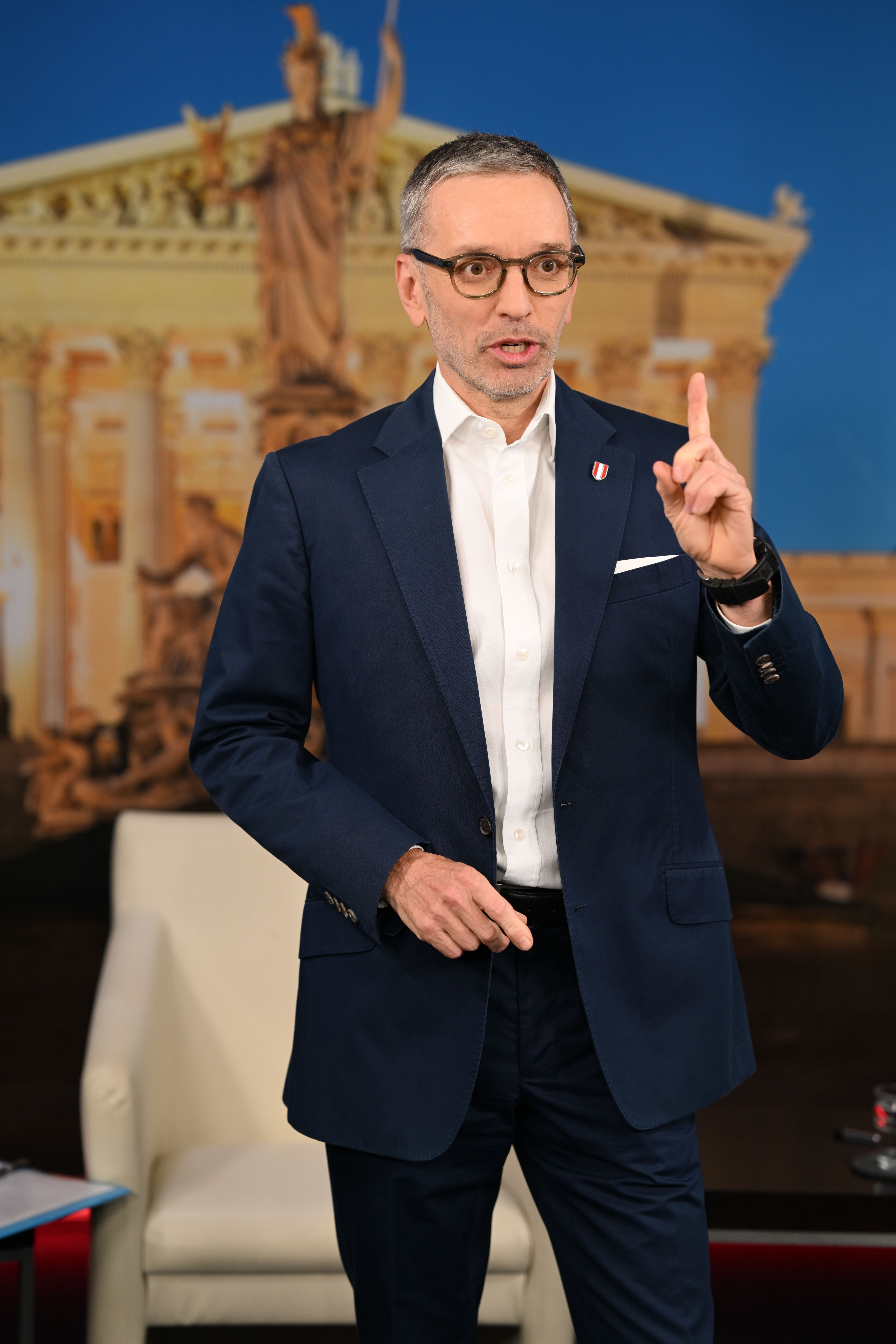 Ein Mann im Anzug spricht und hebt einen Finger, im Hintergrund ein historisches Gebäude.