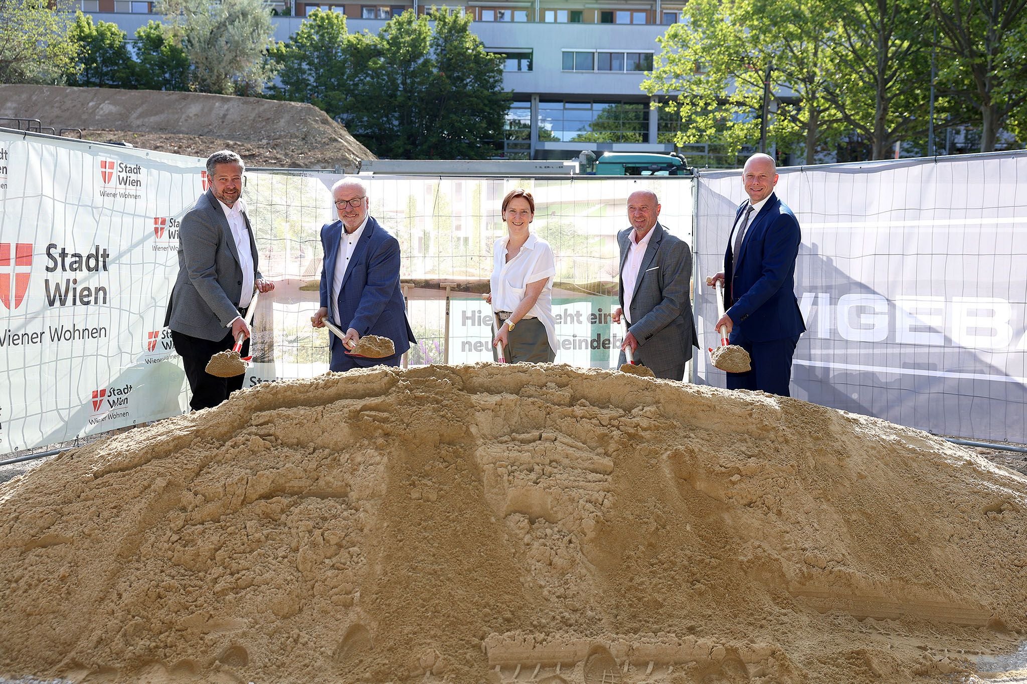 Fünf Personen in Businesskleidung heben Schaufeln Sand bei einem Spatenstich in Wien.