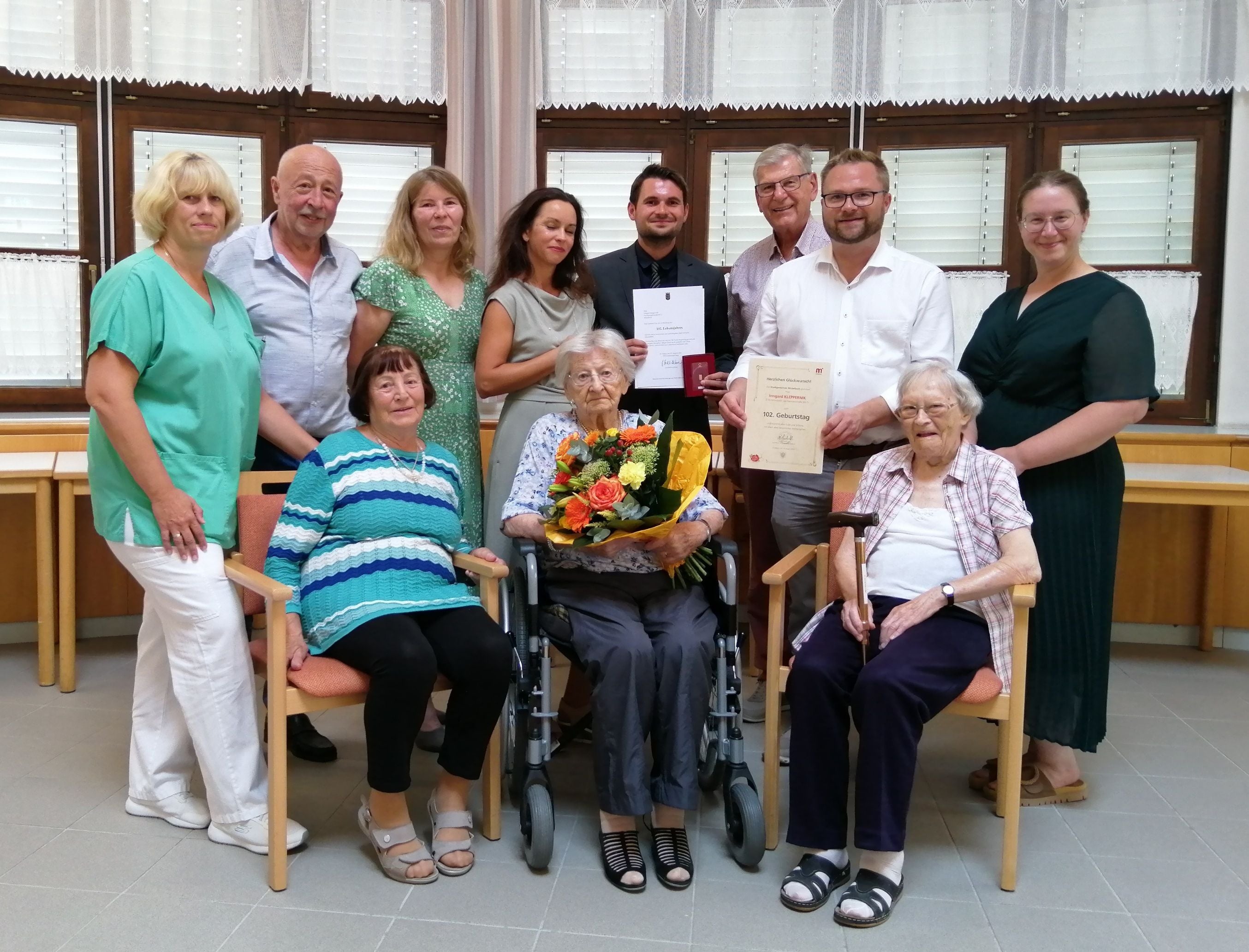 Eine ältere Frau im Rollstuhl erhält zum 102. Geburtstag Blumen, umgeben von Gratulanten.