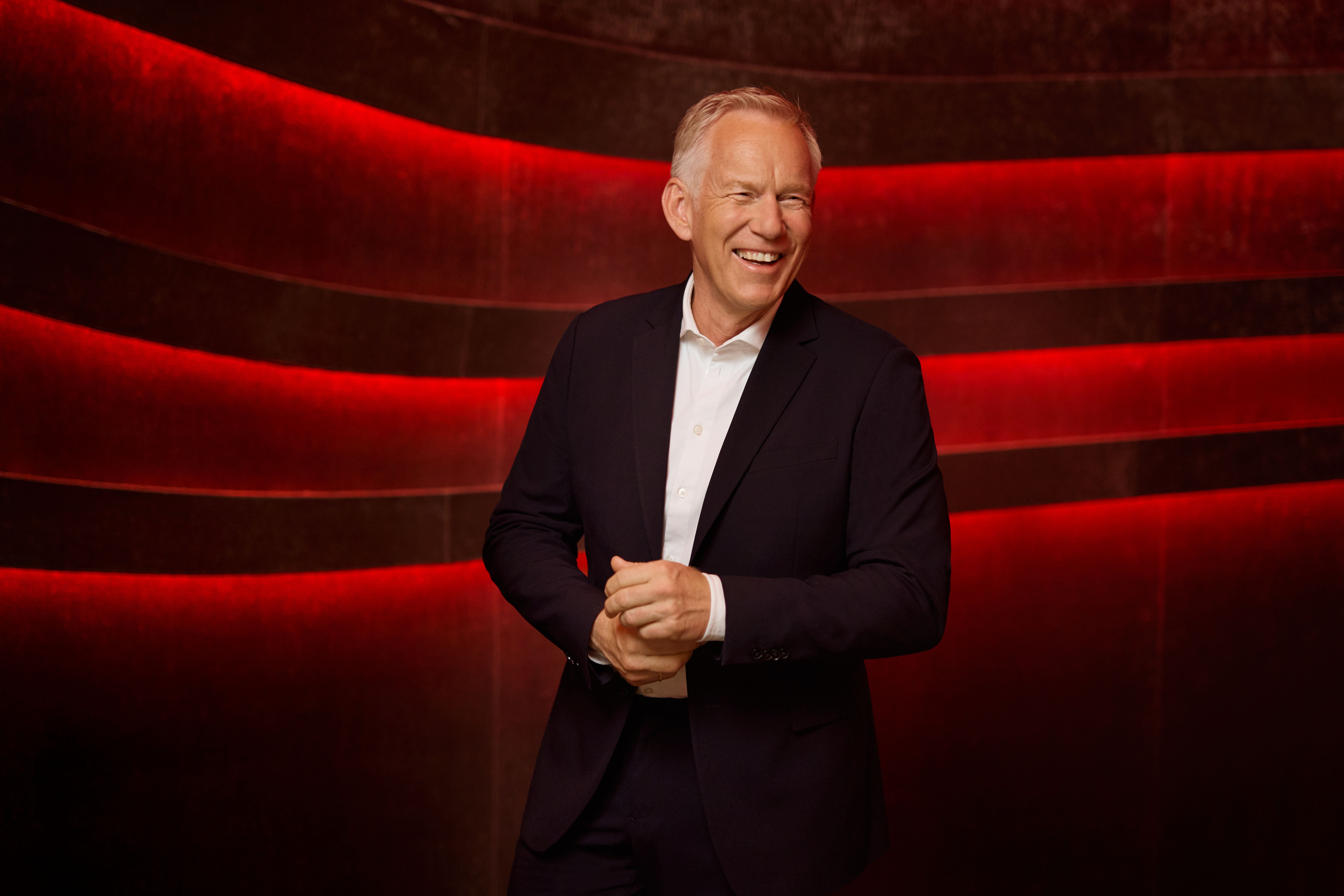 Ein lächelnder Mann im Anzug steht vor einer Wand mit roten Lichtstreifen.