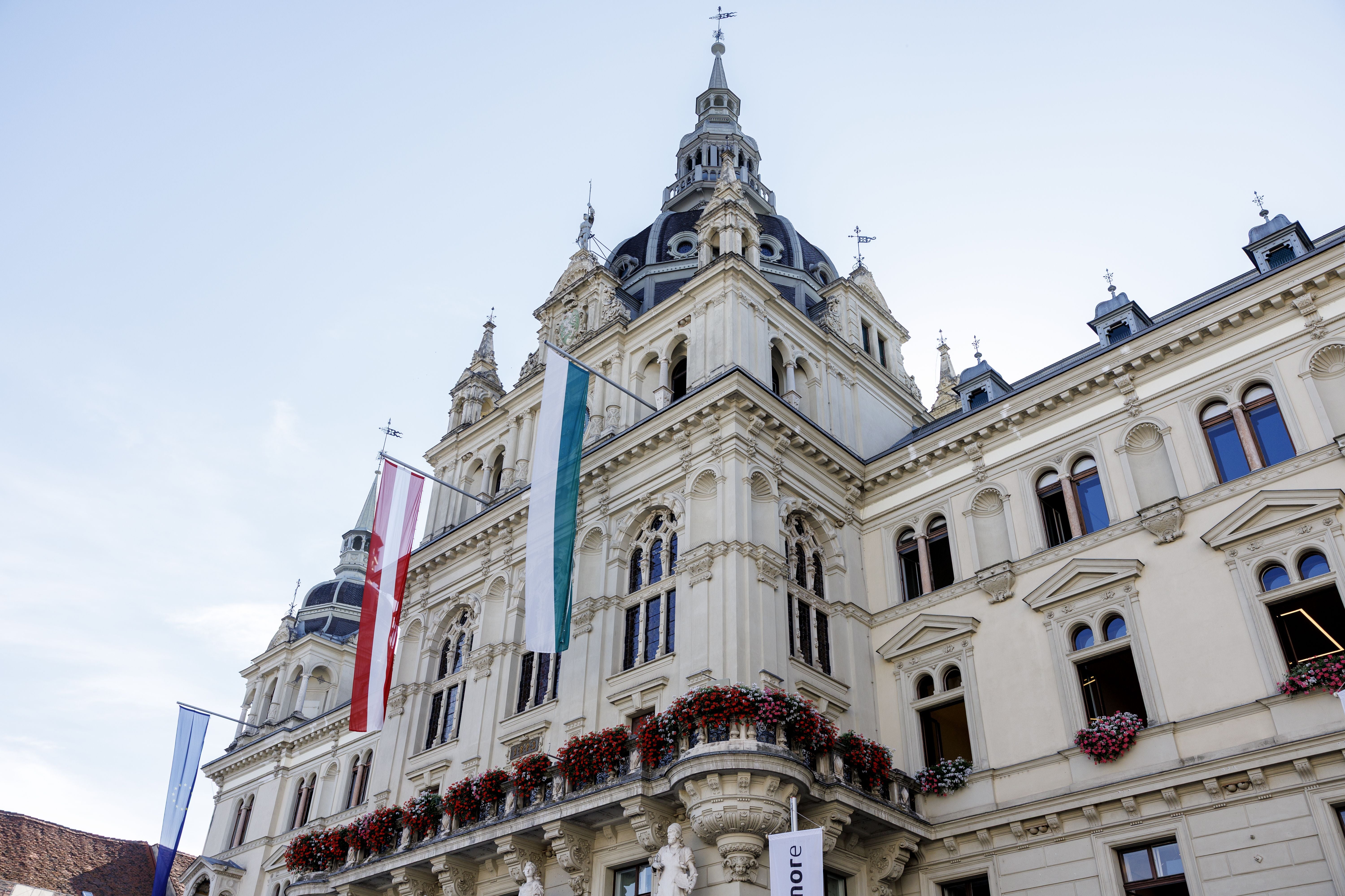 Das Grazer Rathaus mit Blumenbalkonen und wehenden Fahnen vor blauem Himmel.
