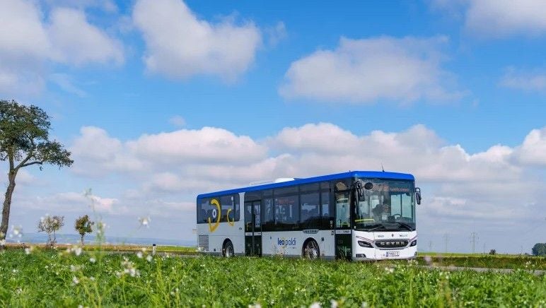 Ein Linienbus mit blauem Dach fährt durch eine grüne Landschaft unter blauem Himmel.