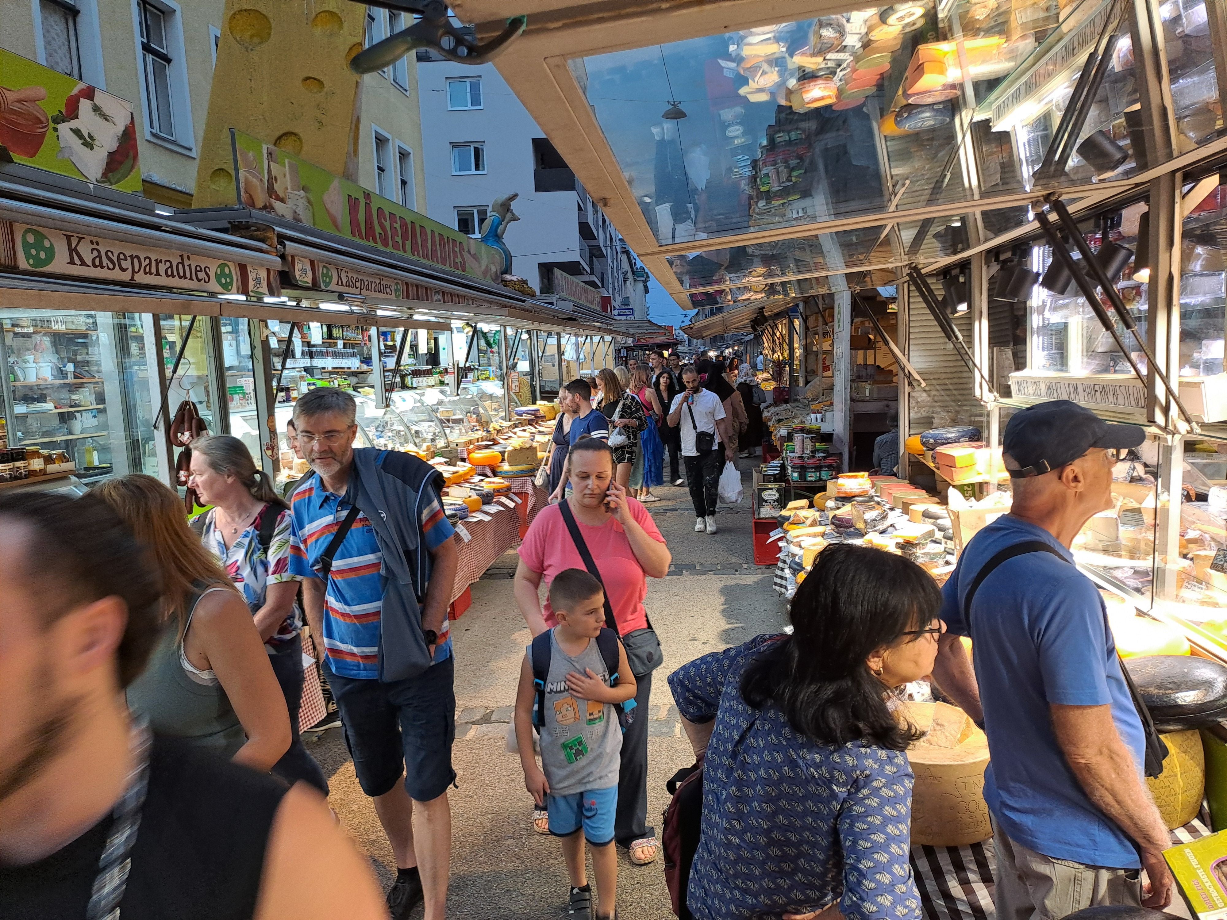 Menschen schlendern durch einen belebten Käsemarkt mit verschiedenen Käseständen.