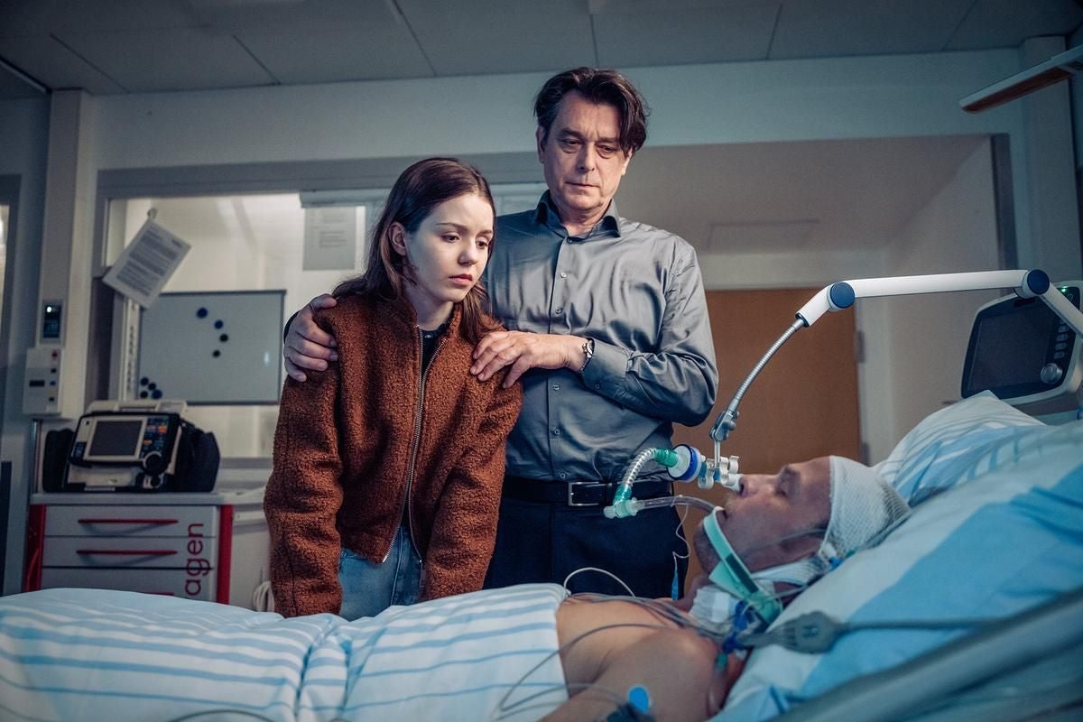 Ein Mann und ein Mädchen stehen traurig am Bett eines beatmeten Patienten im Krankenhaus.