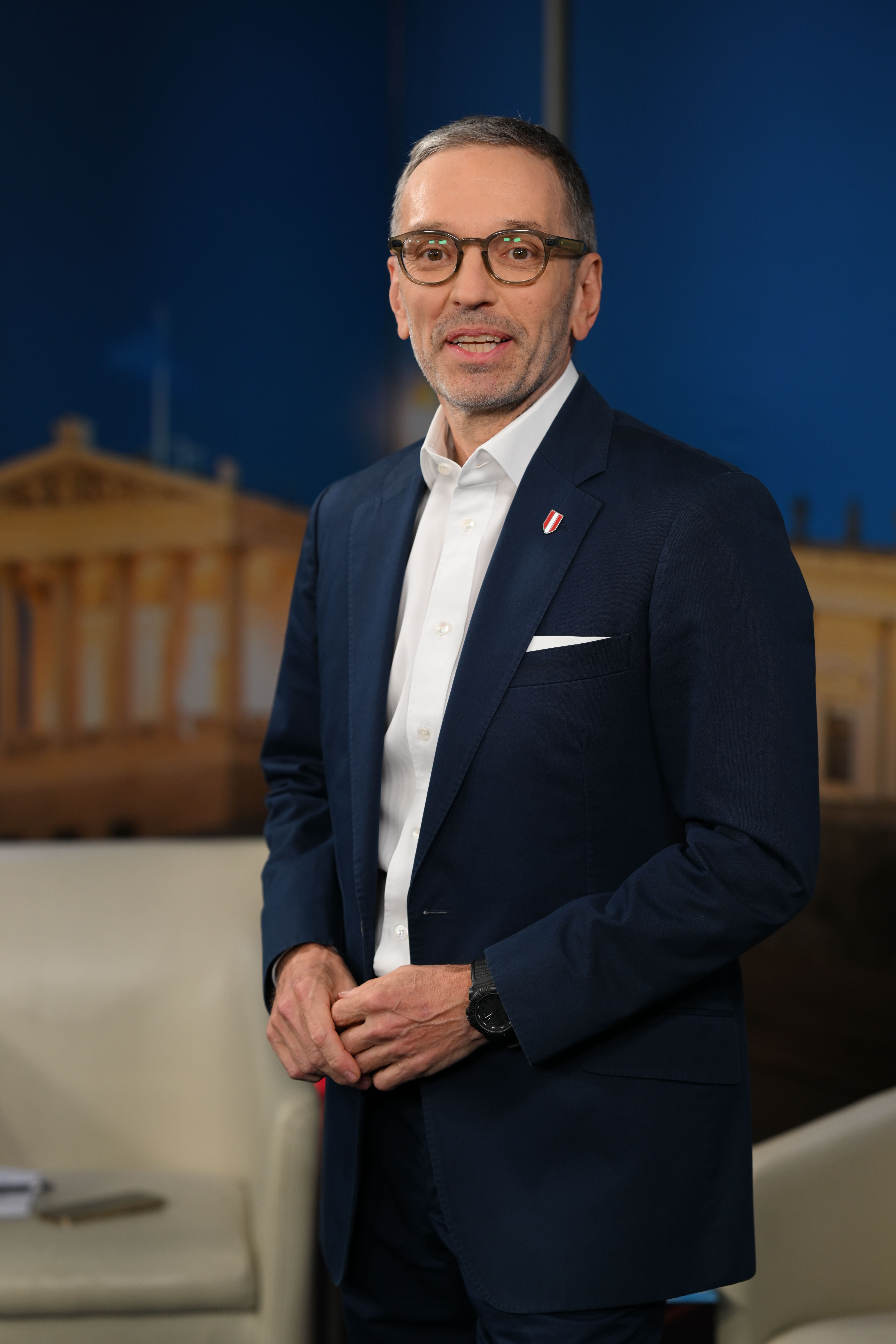 Ein Mann im Anzug mit Brille steht vor einem blauen Hintergrund und schaut in die Kamera.