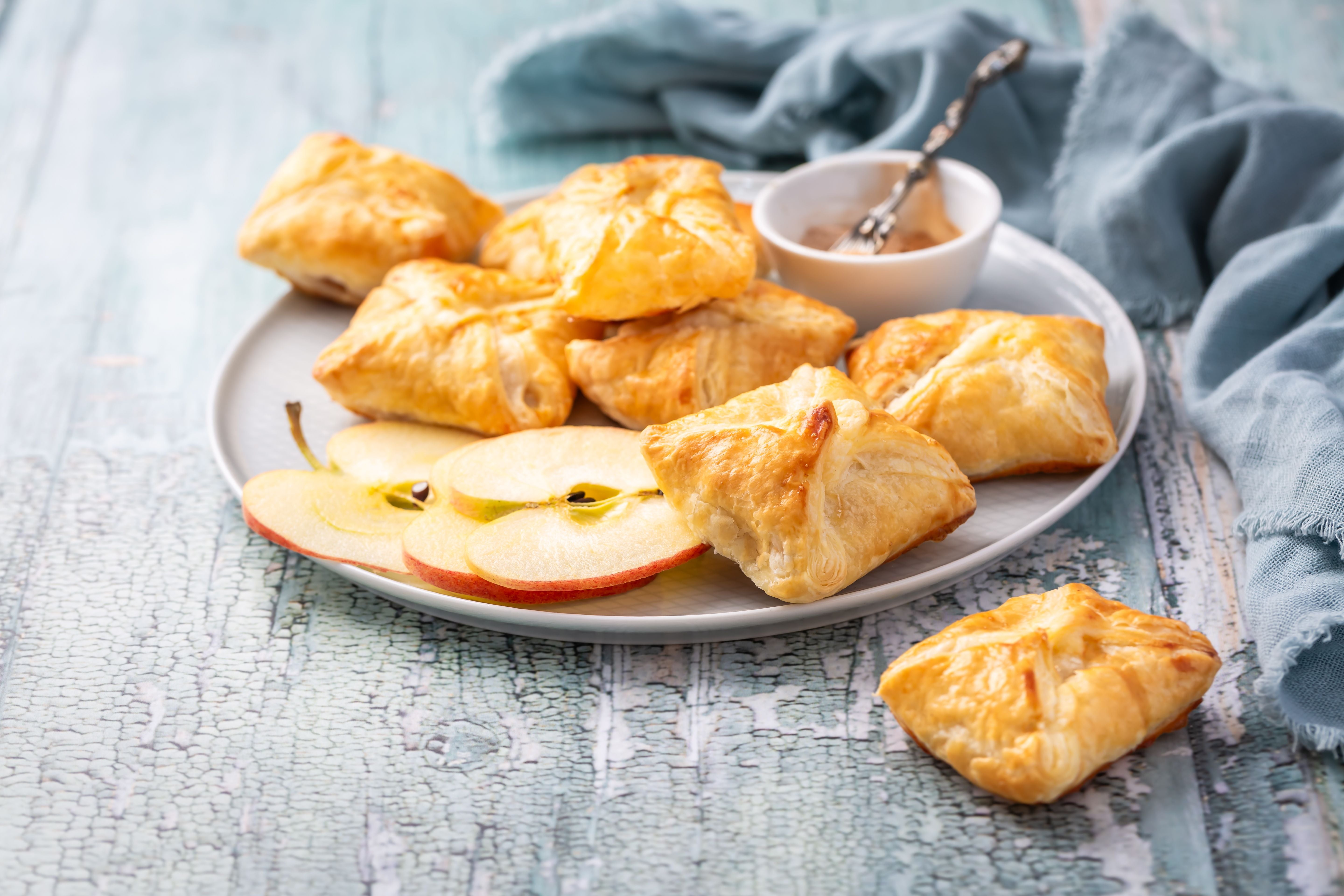 Teller mit selbstgemachten Apfeltaschen aus Blätterteig, Apfelscheiben und einer kleinen Schale Soße.