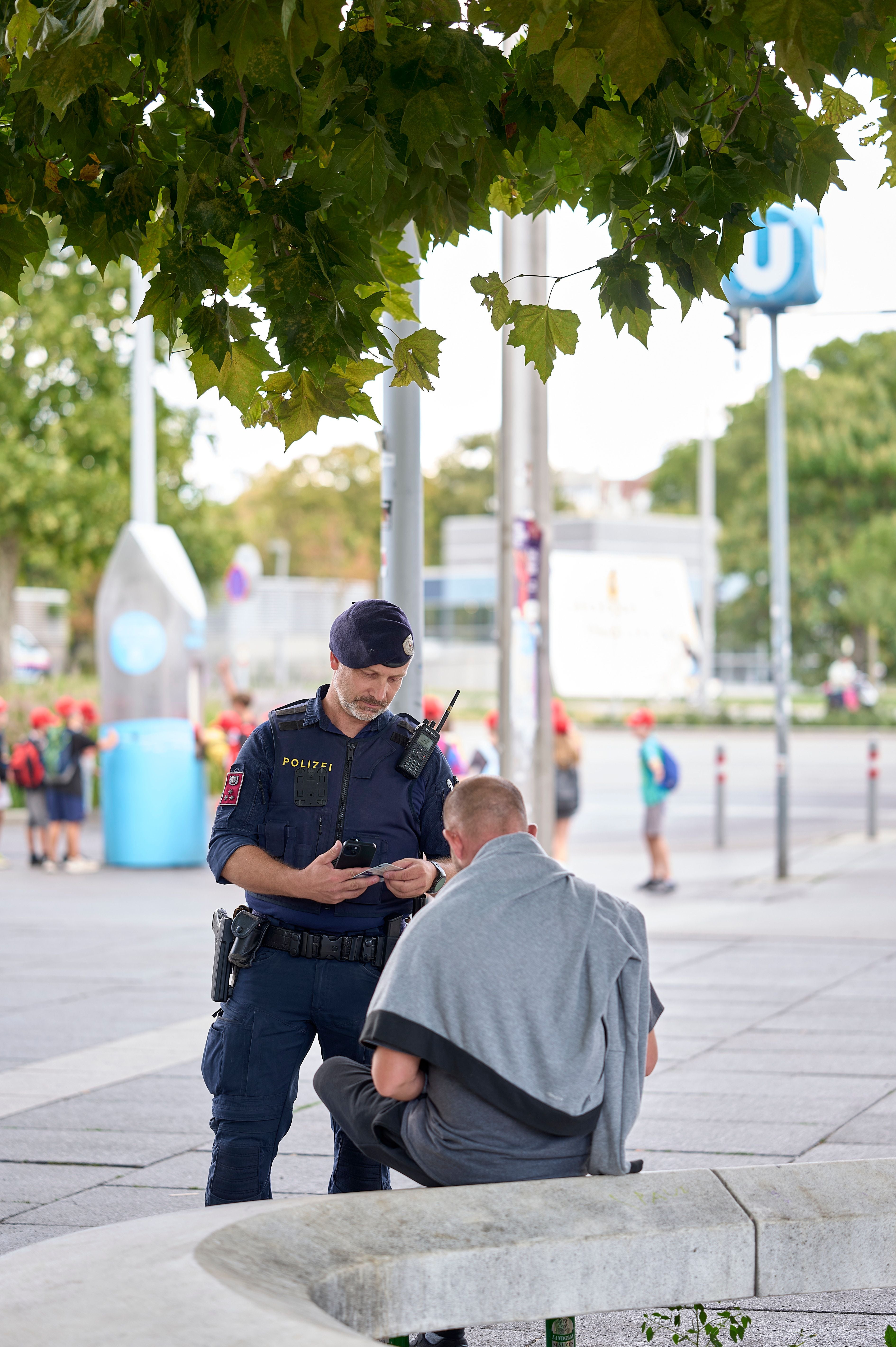 Ein Polizist kontrolliert die Papiere eines sitzenden Mannes im öffentlichen Raum in Wien.
