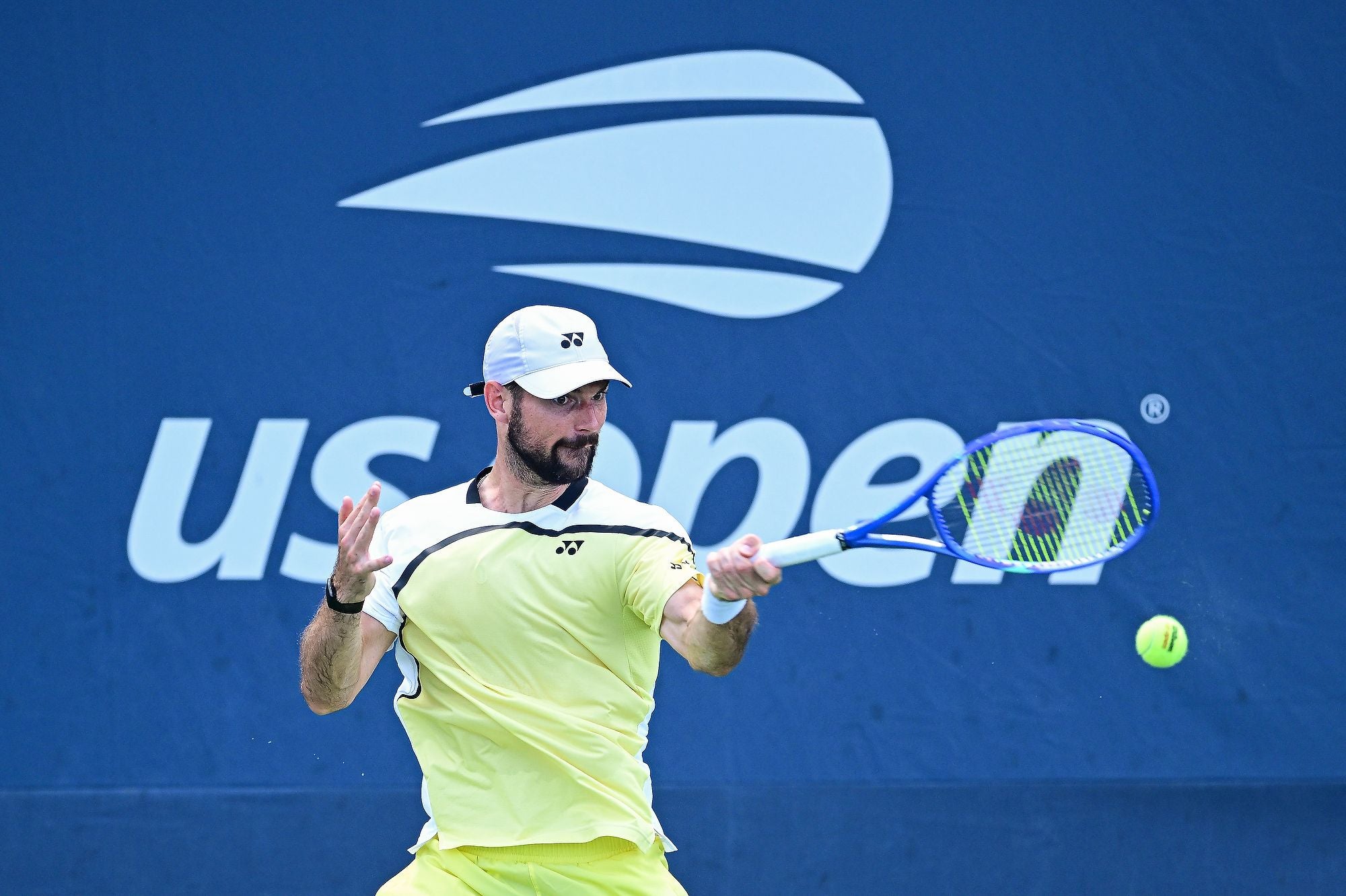 Ein Tennisspieler schlägt den Ball bei den US Open vor blauem Hintergrund mit US-Open-Logo.
