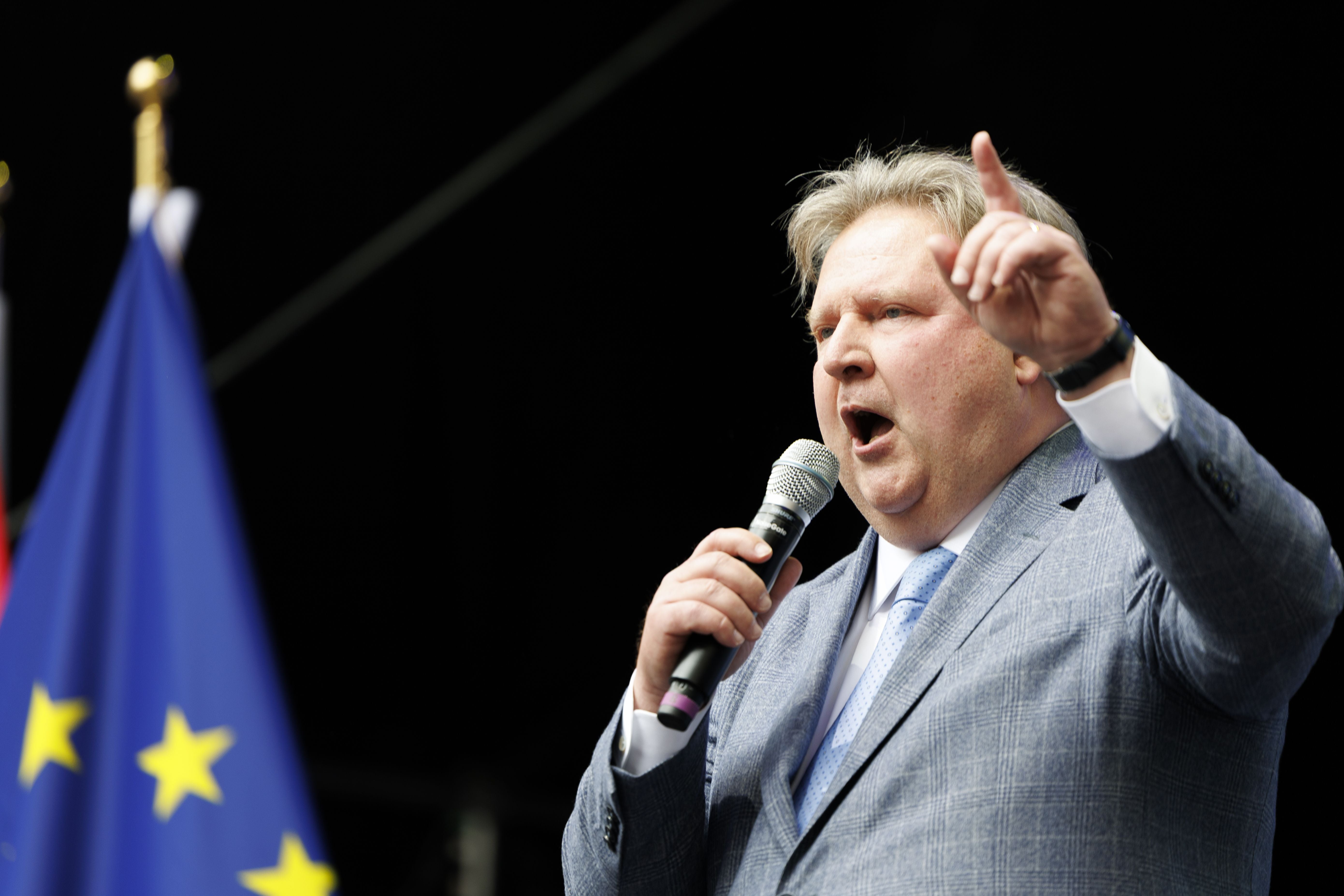 Ein Mann im Anzug spricht mit Mikrofon auf einer Bühne, im Vordergrund eine EU-Flagge.