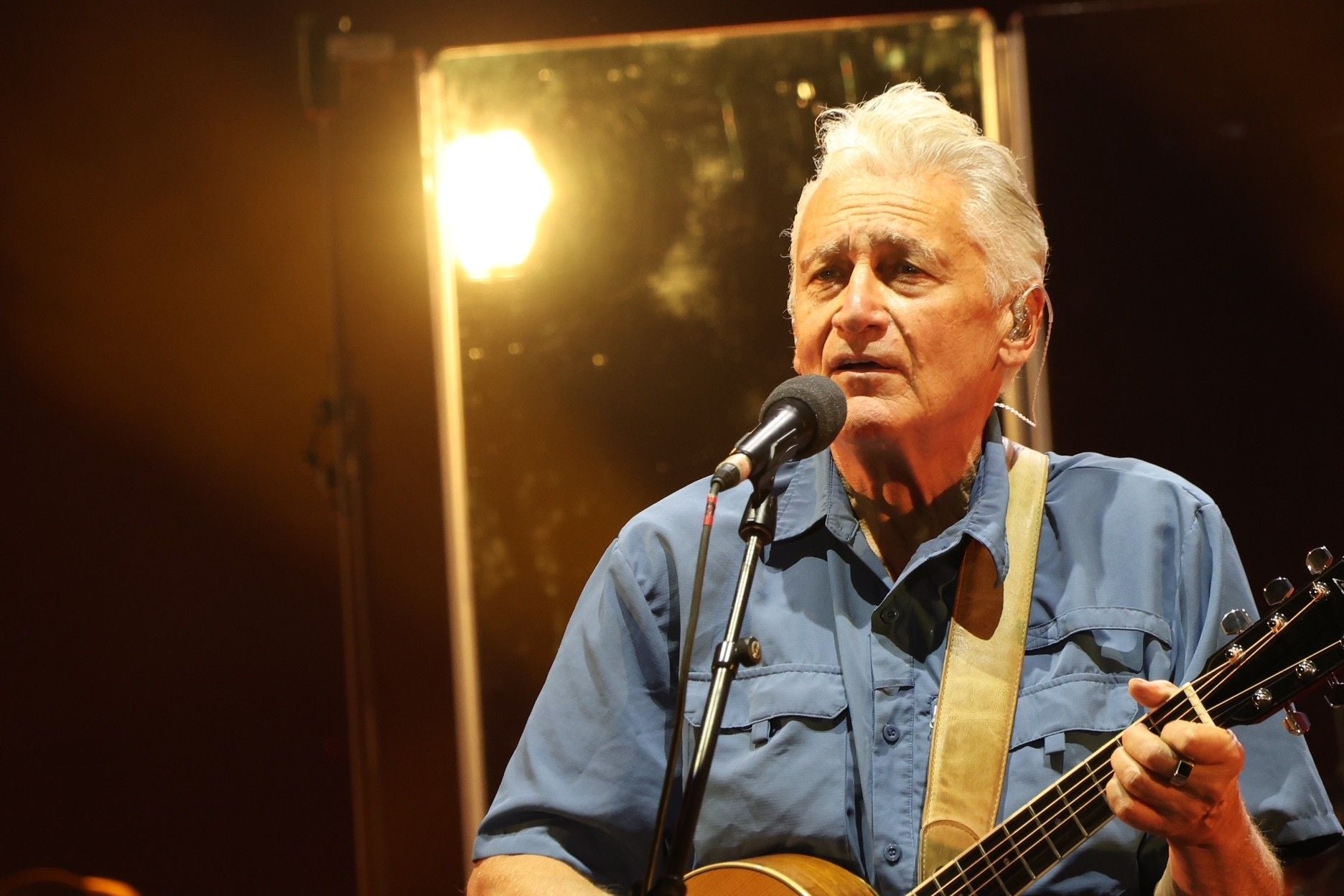 Ein älterer Mann singt und spielt Gitarre auf einer Bühne mit Mikrofon und Bühnenlicht.